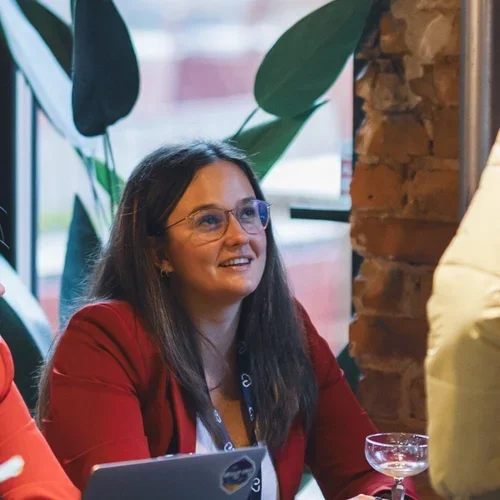 A woman with long dark hair, glasses, and a red jacket smiling at a social event, sitting by a table with a drink in a glass.