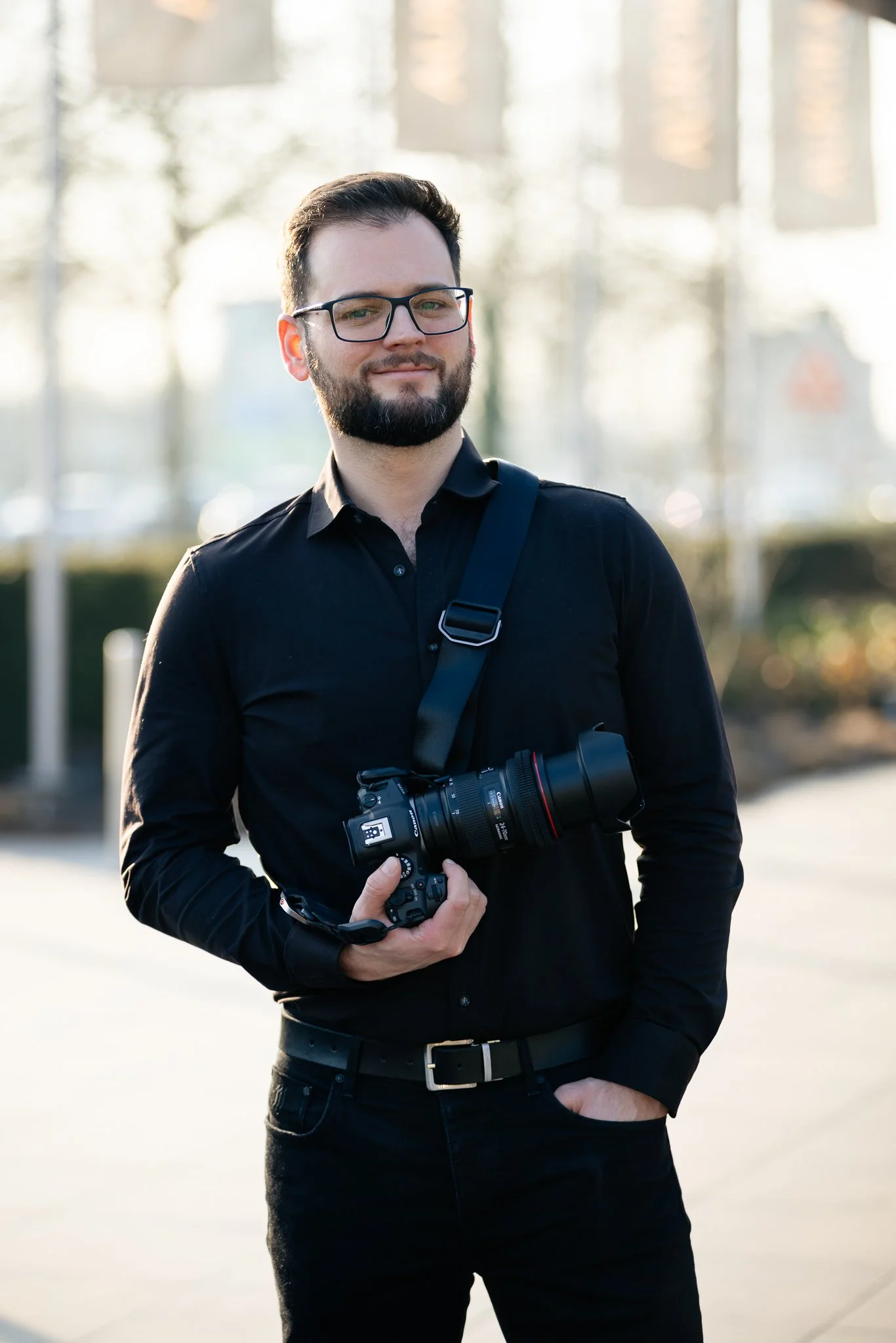 A man with glasses and a beard, dressed in black, holding a professional camera. He is outdoors with blurred trees and buildings in the background.