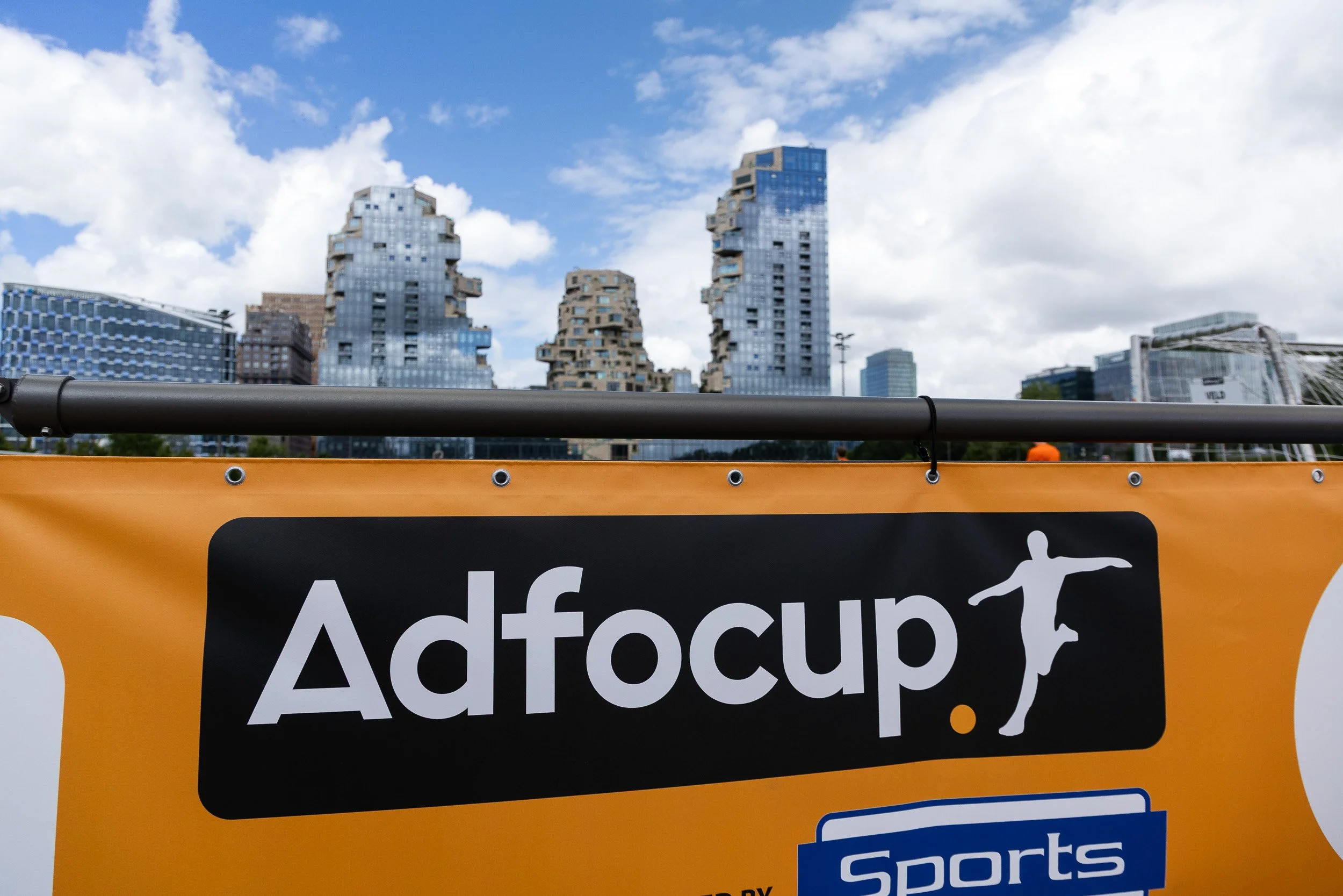 Adfocup 2026 orange branded pitch hoarding with Amsterdam Zuidas skyline in background — corporate event photography Amsterdam Ballo Studio