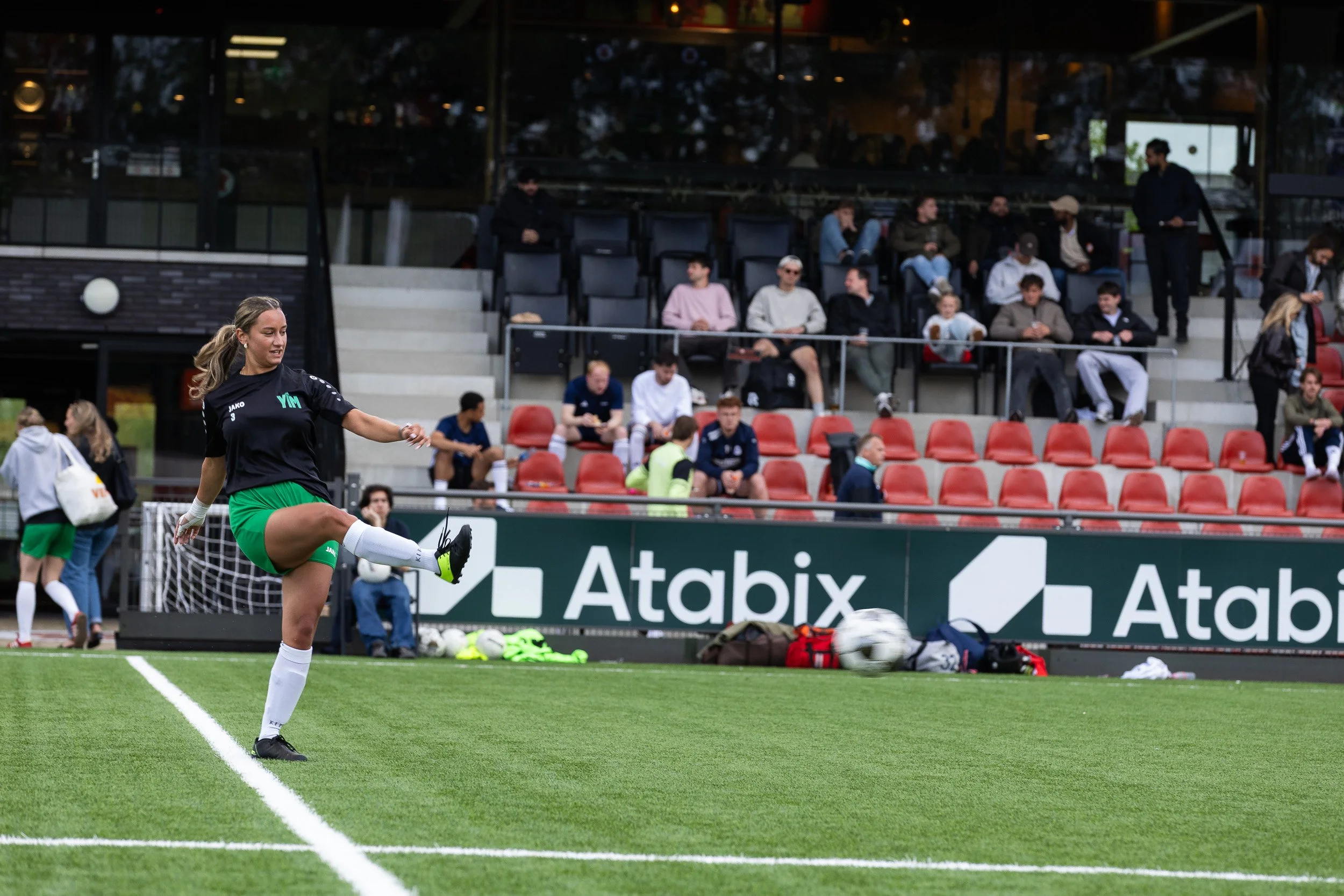 Female player in green and black kit kicking ball during Adfocup 2026 match at Amsterdamsche Football Club — event photographer Amsterdam Ballo Studio