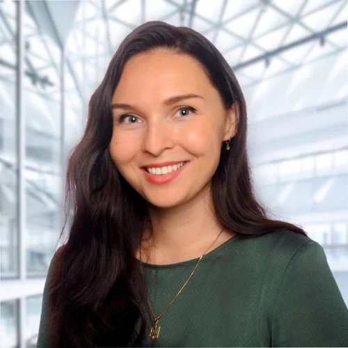 Portrait of a young woman with long dark hair, smiling, wearing a green top and gold necklace, in a modern glass building interior.