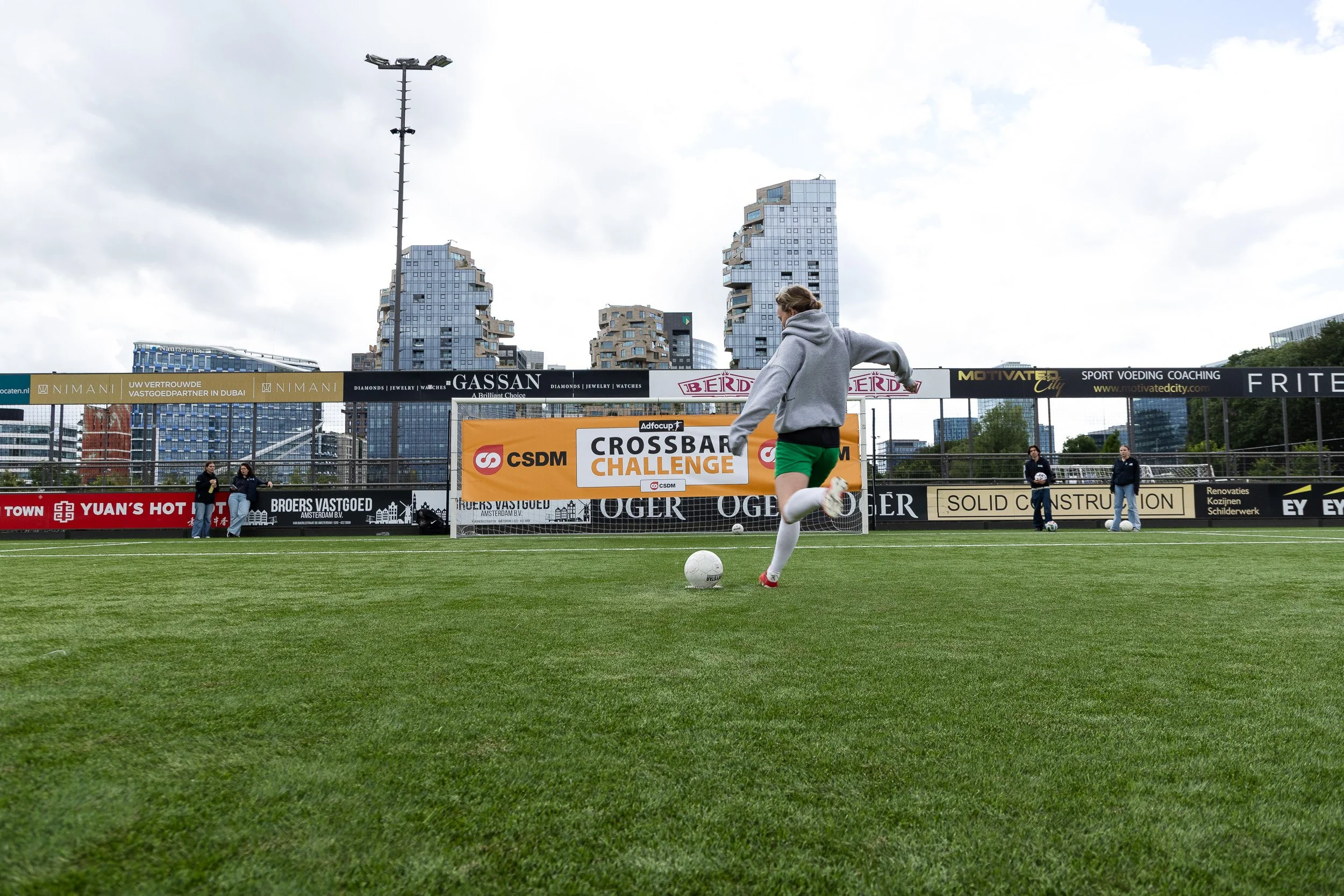 Player in orange kit kicking football at Adfocup 2026 Crossbar Challenge on pitch with Amsterdam skyline — event photography Amsterdam Ballo Studio