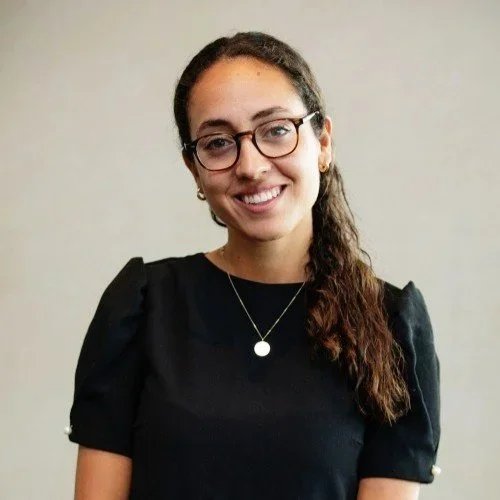 A young woman with glasses and long, curly brown hair smiling in front of a plain wall.