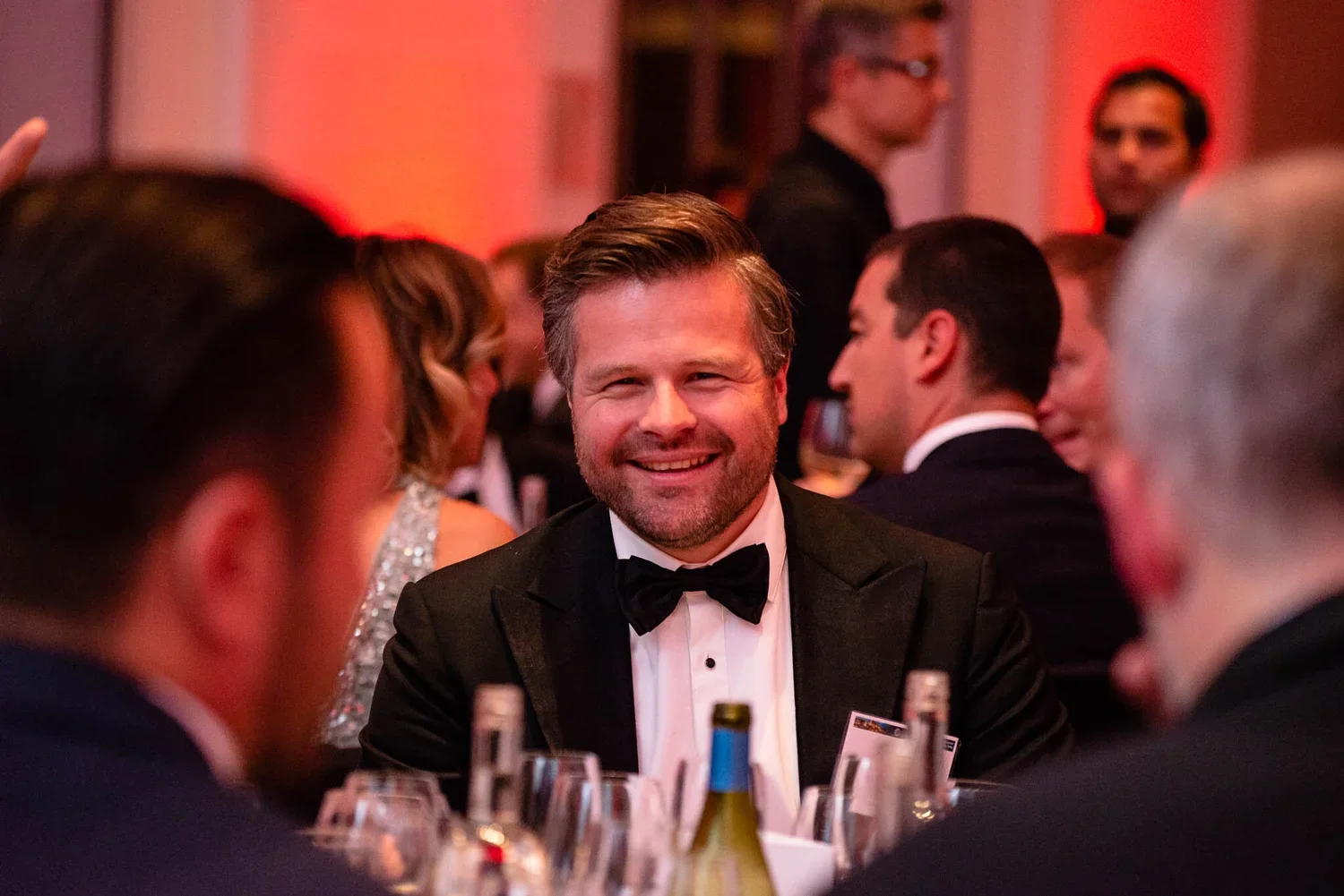 A man in a tuxedo smiling at a formal event, surrounded by other guests in formal attire, at a dinner or banquet with a warm, reddish lighting.