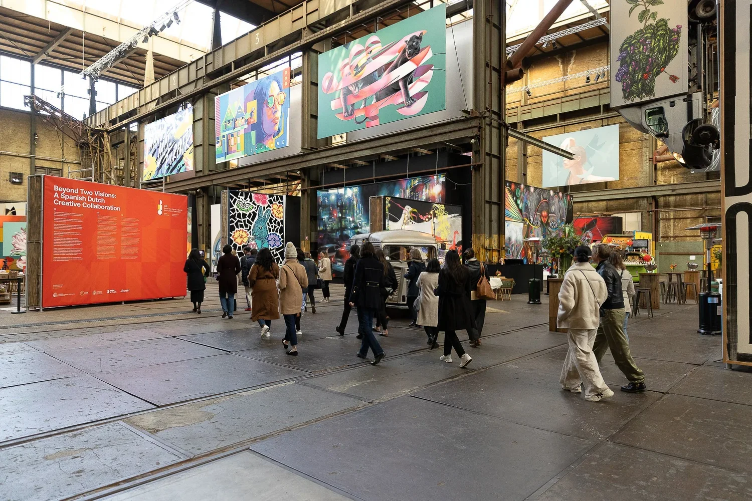 People walking inside an industrial-style art gallery or exhibition space with large colorful murals and artworks displayed on the walls and overhead.