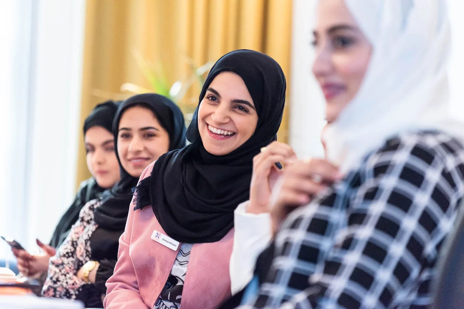 Four women wearing hijabs sitting at a conference table, with the woman in the pink blazer smiling and engaging in conversation.