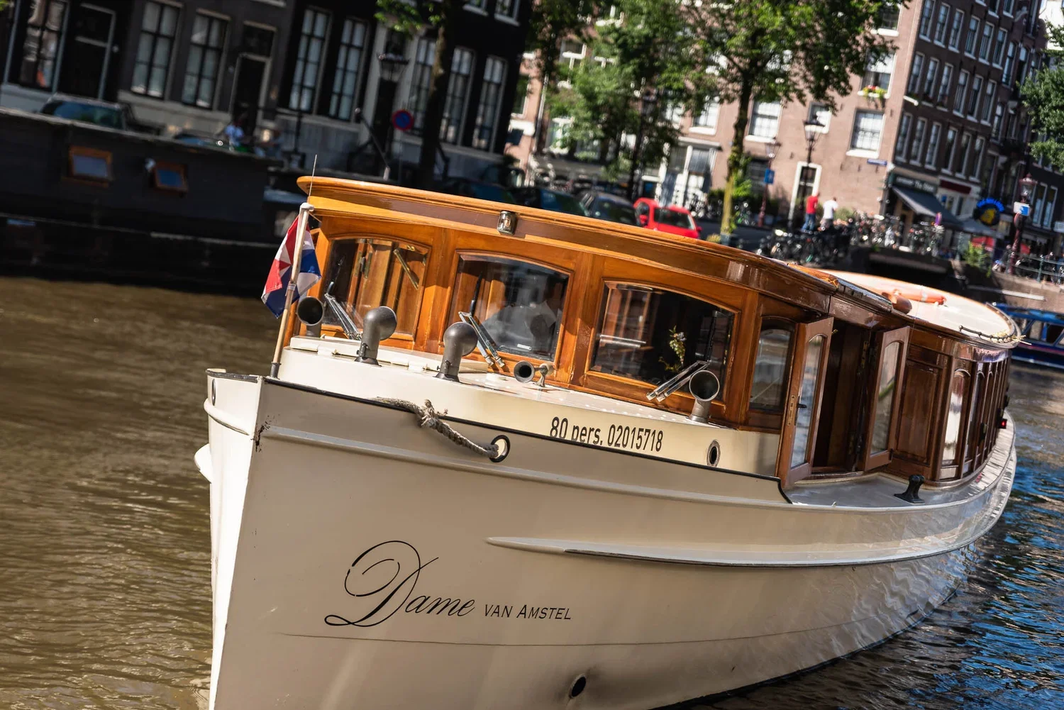 A wooden-hulled boat named Dame Van Amstel sailing on a canal with city buildings and trees in the background.