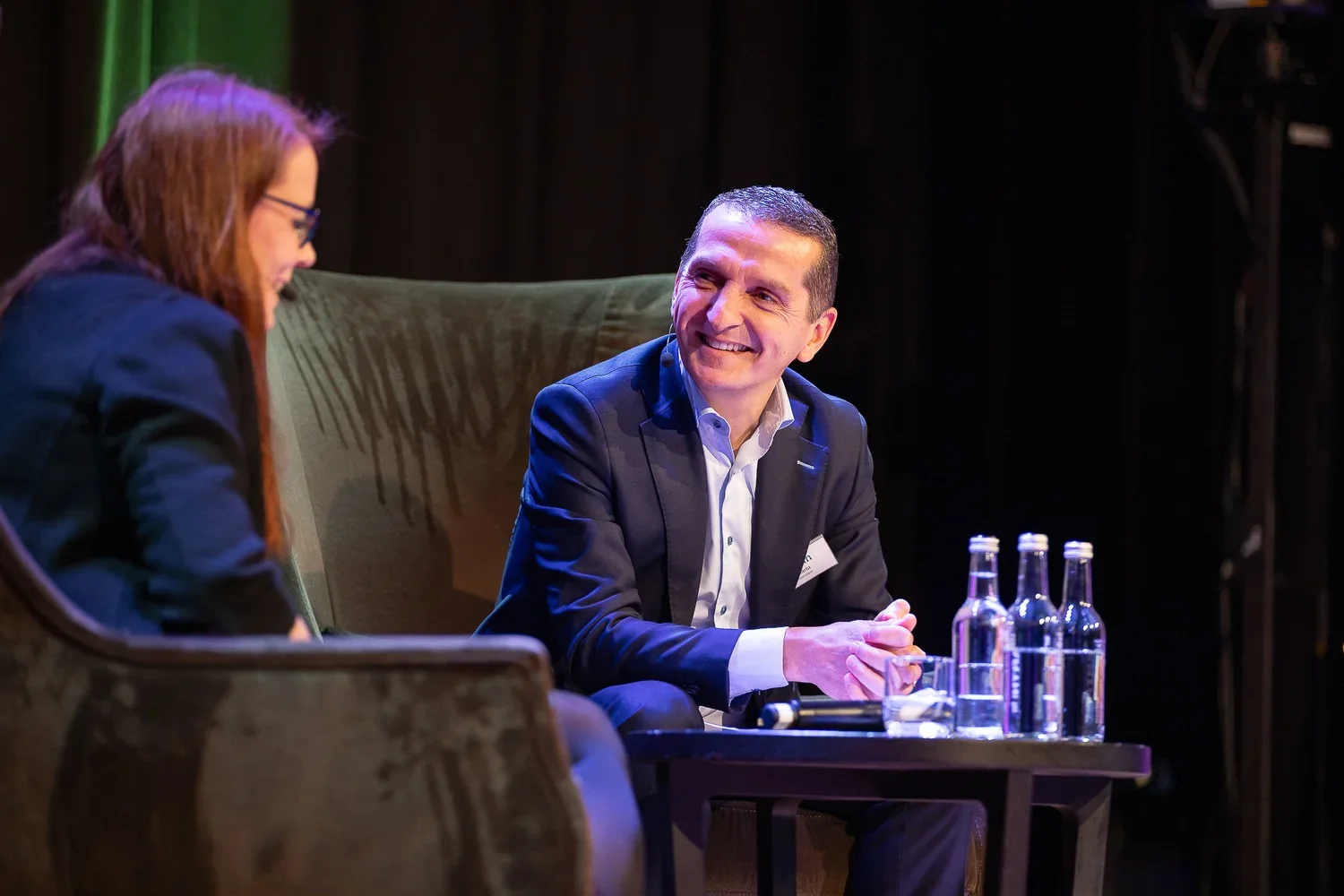 Two people sitting during a discussion on stage, with one smiling and leaning forward, and the other minimally visible, with a table of water bottles between them.