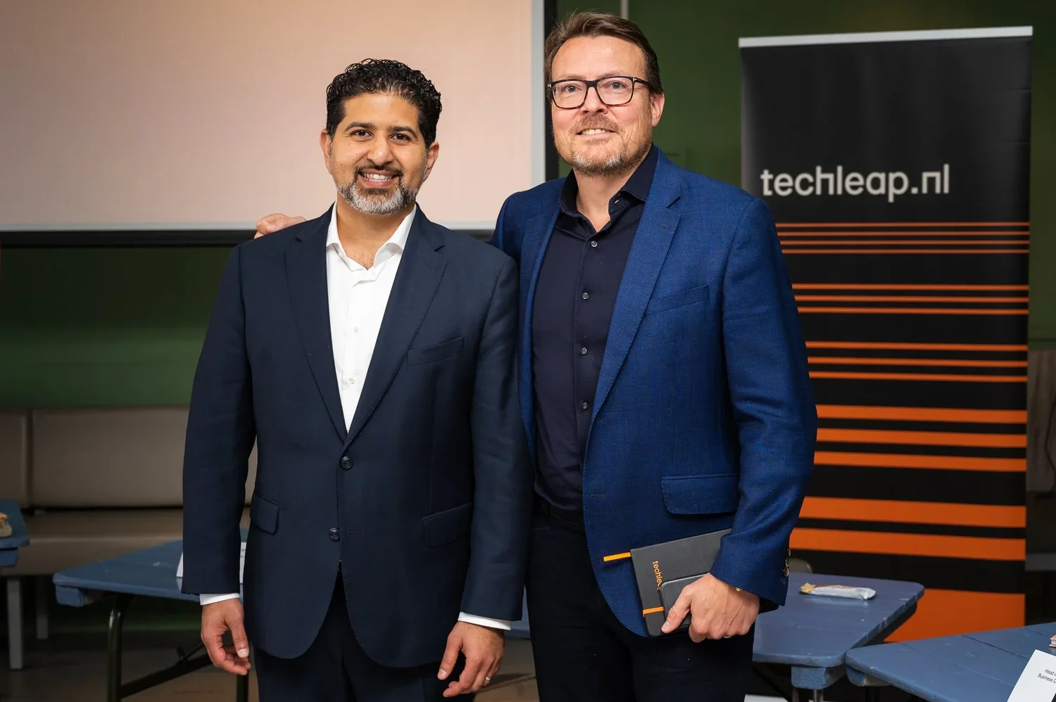 Two men in suits smiling and posing for a photo at a professional event. One holds a notebook, with a tech-related banner in the background.