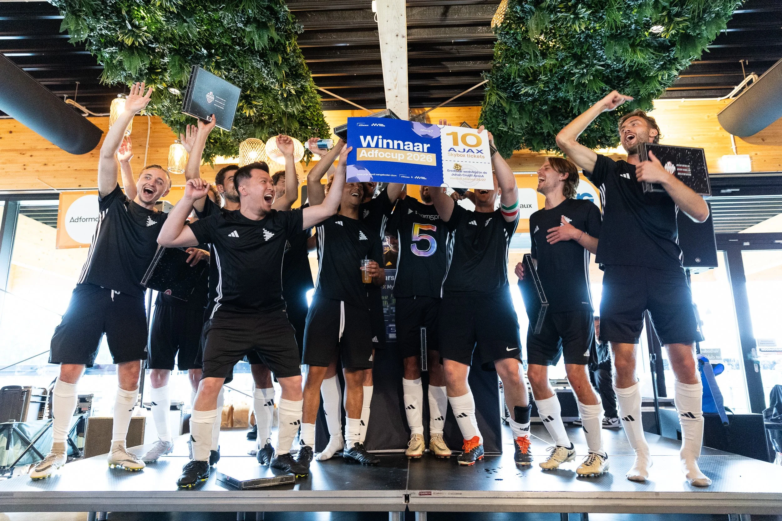 Winning team in black kit celebrating on stage with Adfocup 2026 winners banner and trophy at prize ceremony Amsterdam — event photography Amsterdam Ballo Studio