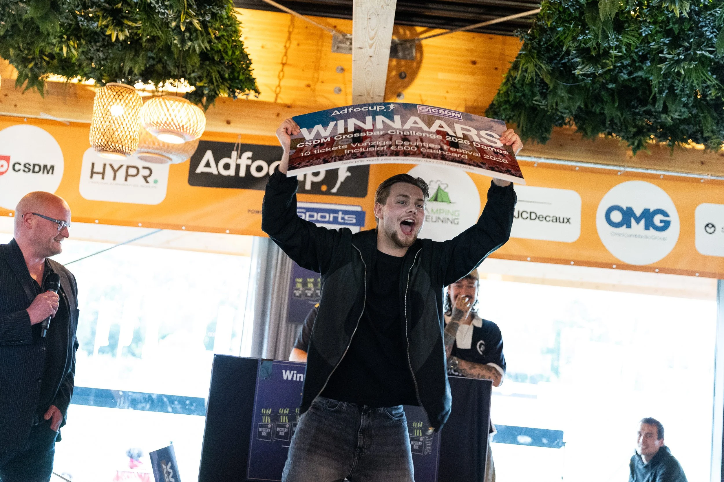 Jubilant winner raising CSDM Crossbar Challenge banner above head on stage at Adfocup 2026 prize ceremony with sponsor backdrop Amsterdam — event photographer Amsterdam Ballo Studio