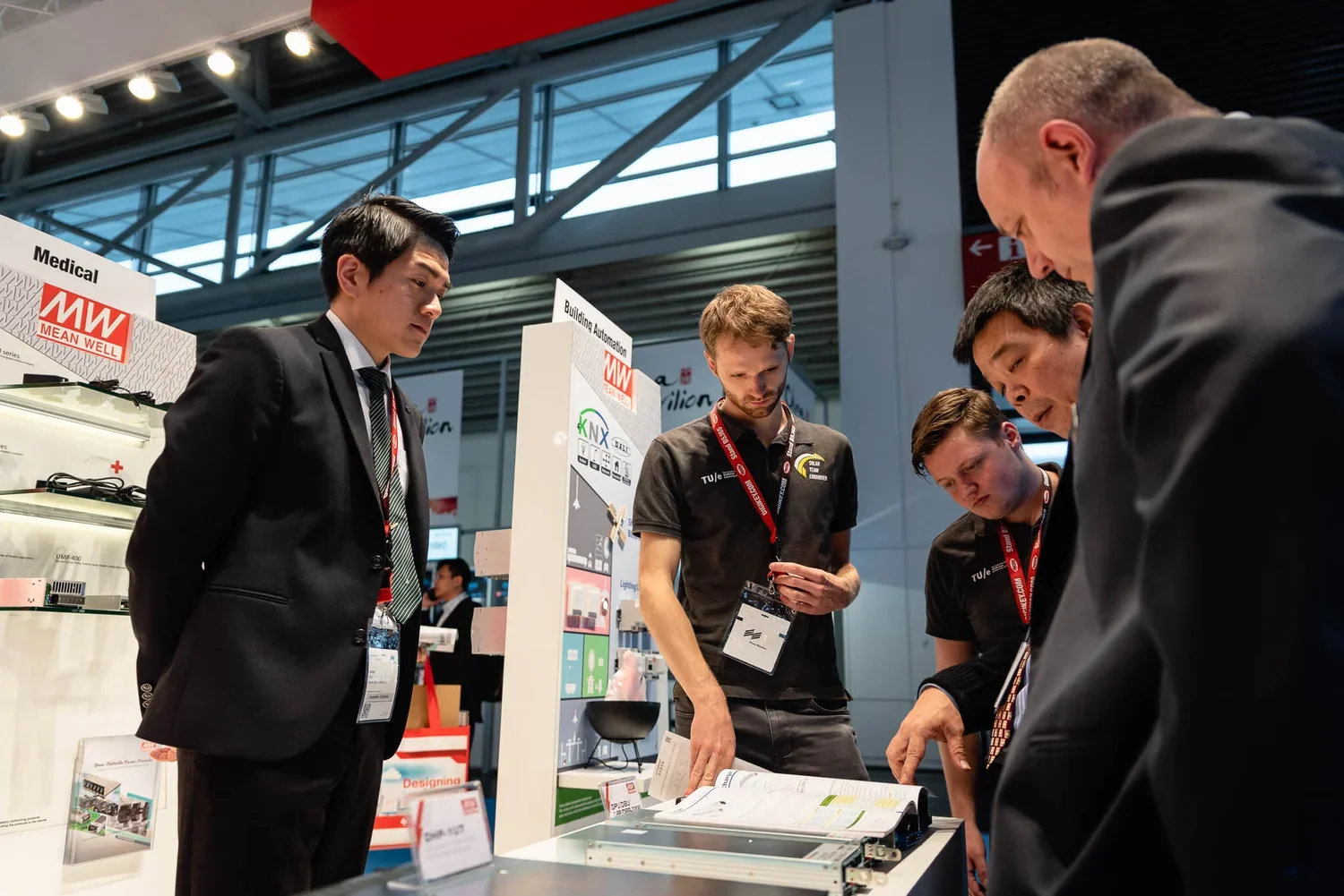 Group of five men at a trade show booth looking at technical documents and product display, with a large banner and exhibition hall background.