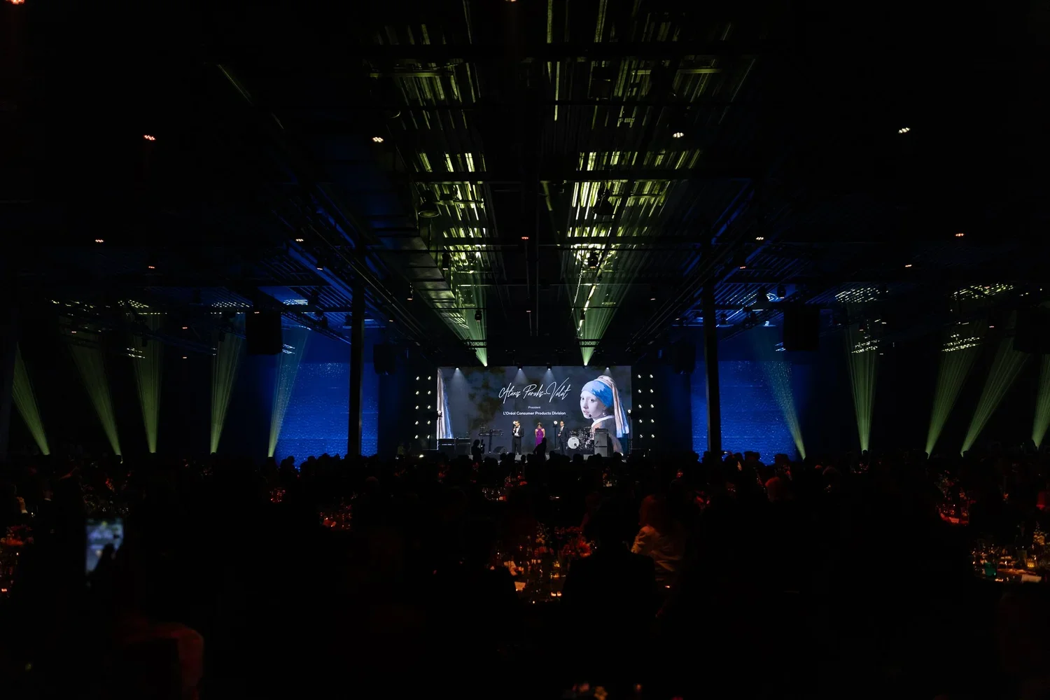 Large conference or awards ceremony with a stage featuring a projection of a girl wearing a blue headband and the text 'Office Parady Awards'. Audience seated at round tables in a dimly lit room with green and blue lighting and spotlights.