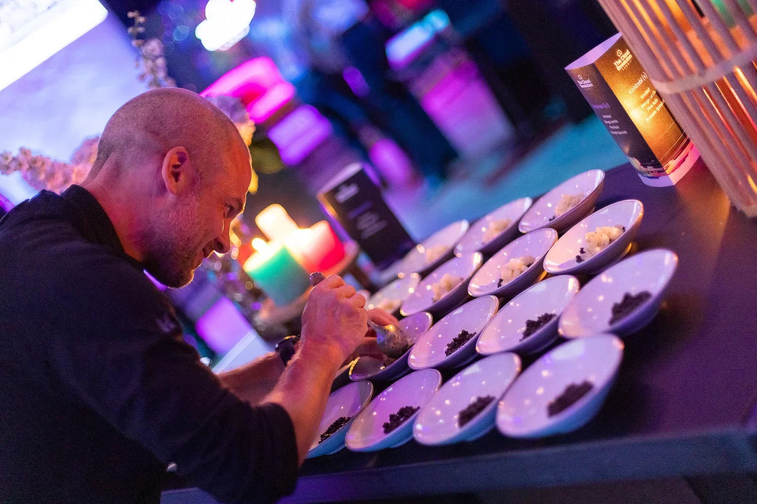 A man with a beard is seasoning bowls of food on a kitchen counter with colorful, dim lighting in the background