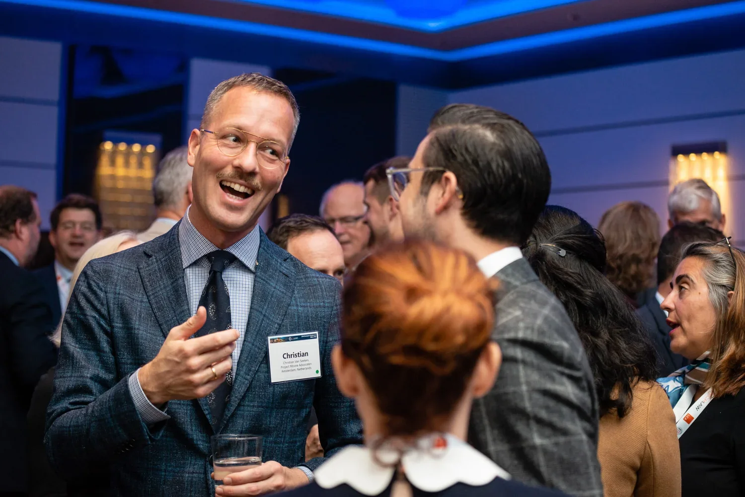 Group of business professionals at a networking event, engaging in conversation, with a man named Christian smiling and holding a drink.