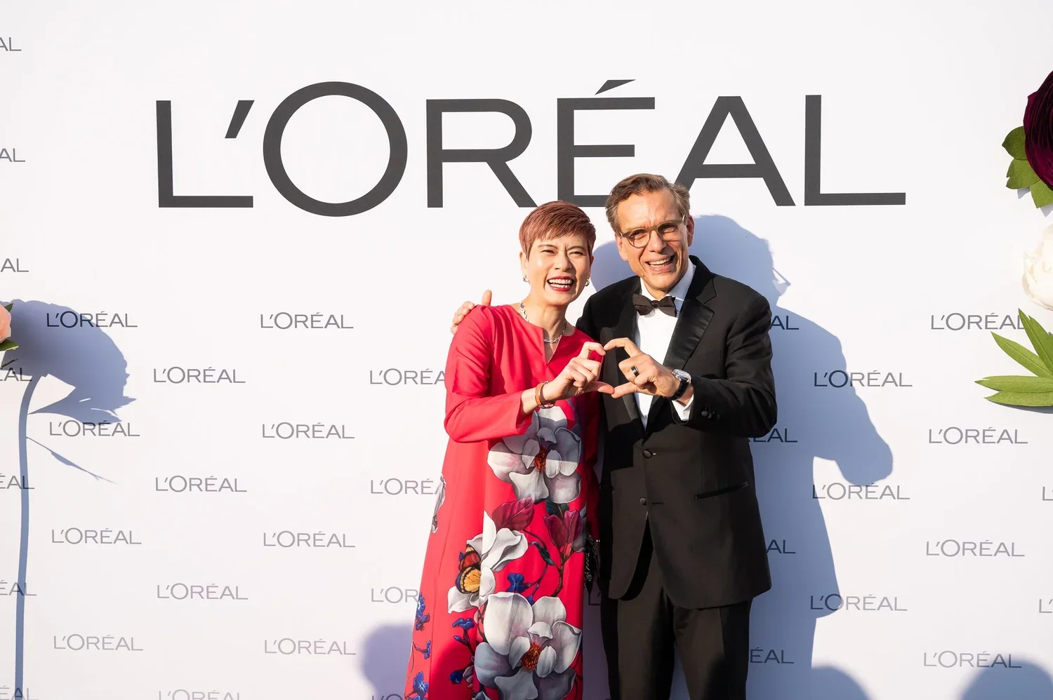 Two people smiling and forming a heart shape with their hands at a L'Oréal event, standing in front of a white background with the L'Oréal logo and floral decorations.