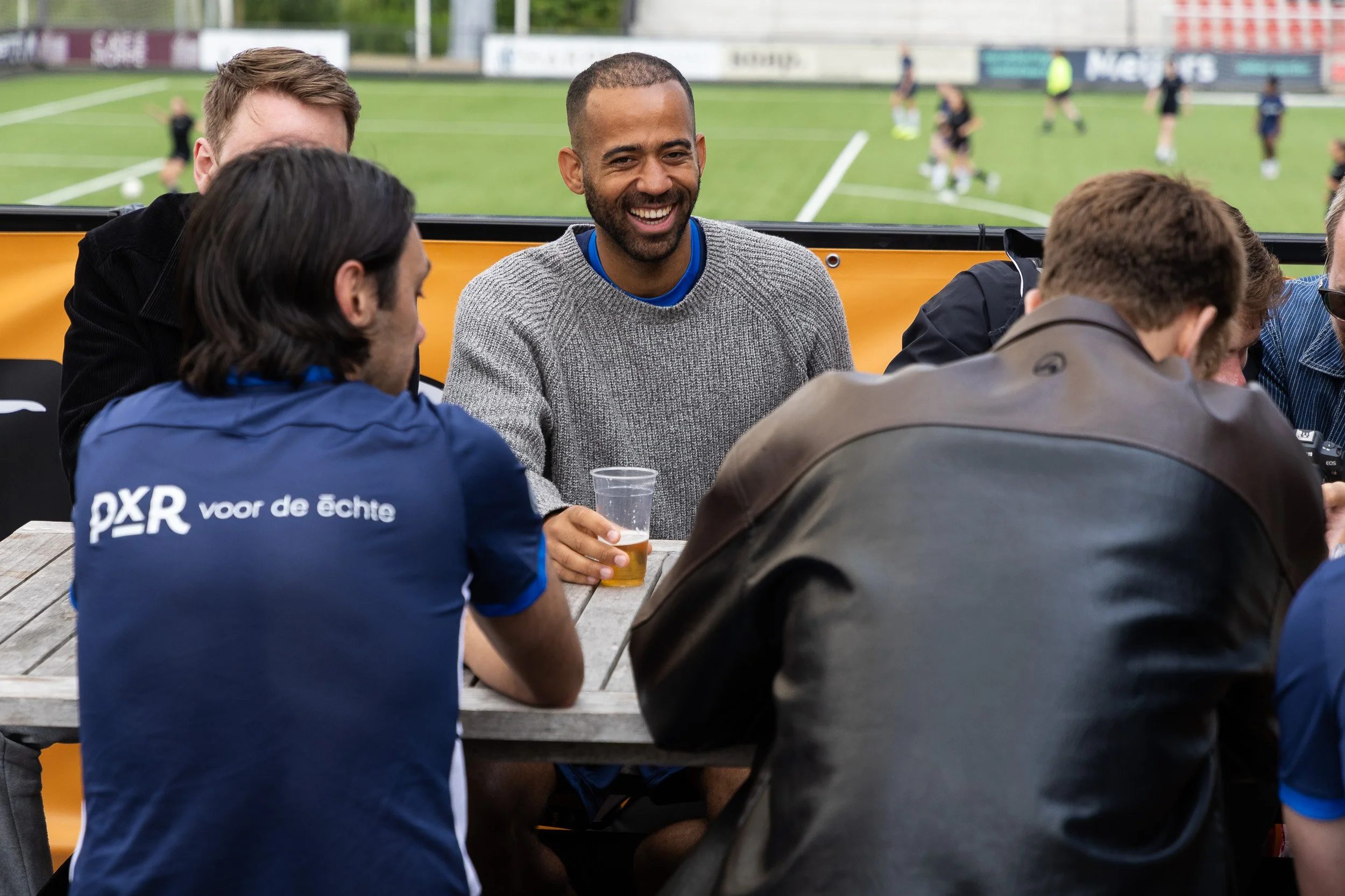 Colleagues networking and laughing with drinks at Adfocup 2026 with football pitch in background Amsterdam — event photography Amsterdam Ballo Studio