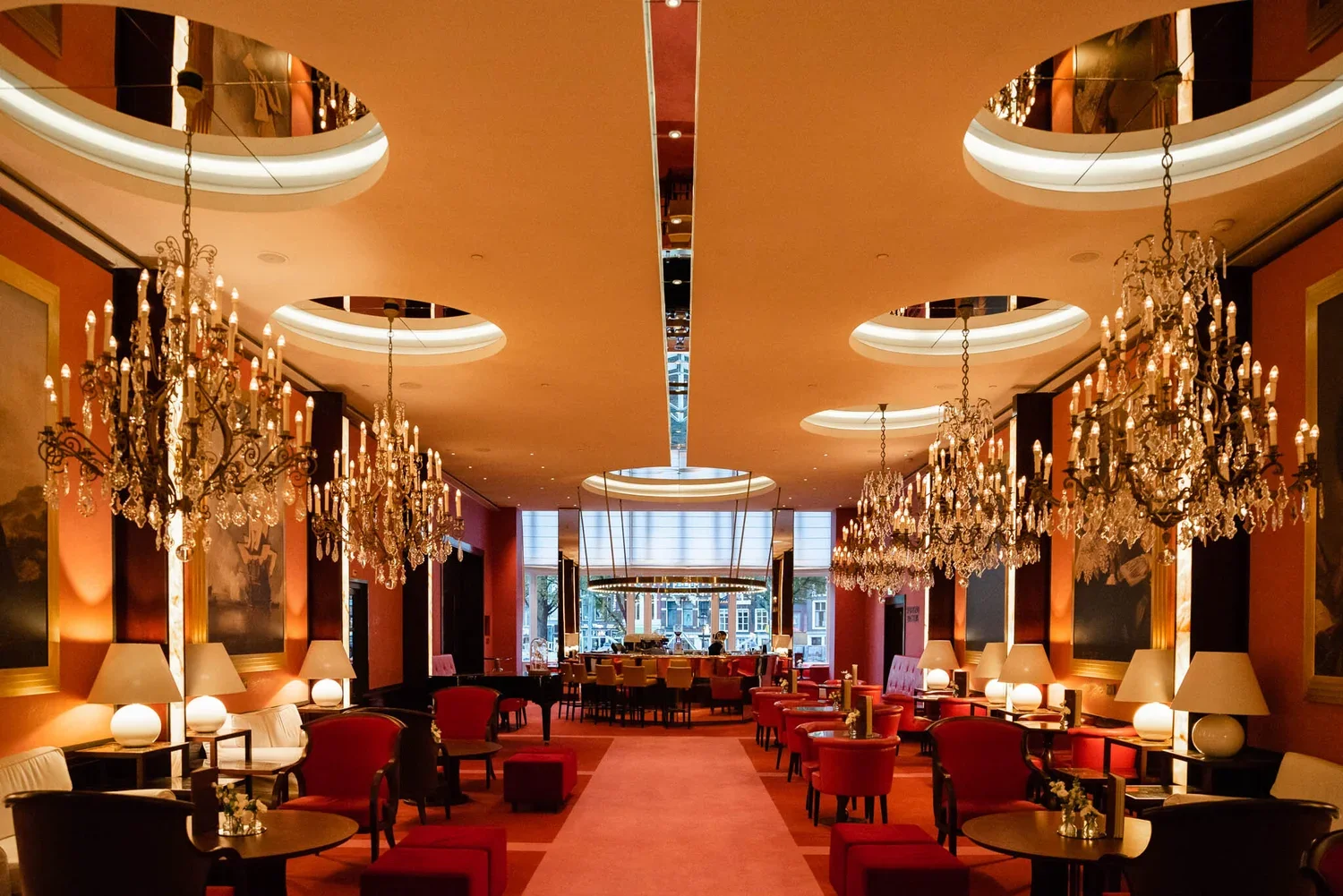 Elegant hotel lounge with chandeliers, red and black chairs, round tables, and lamps, with large windows showing a cityscape outside.