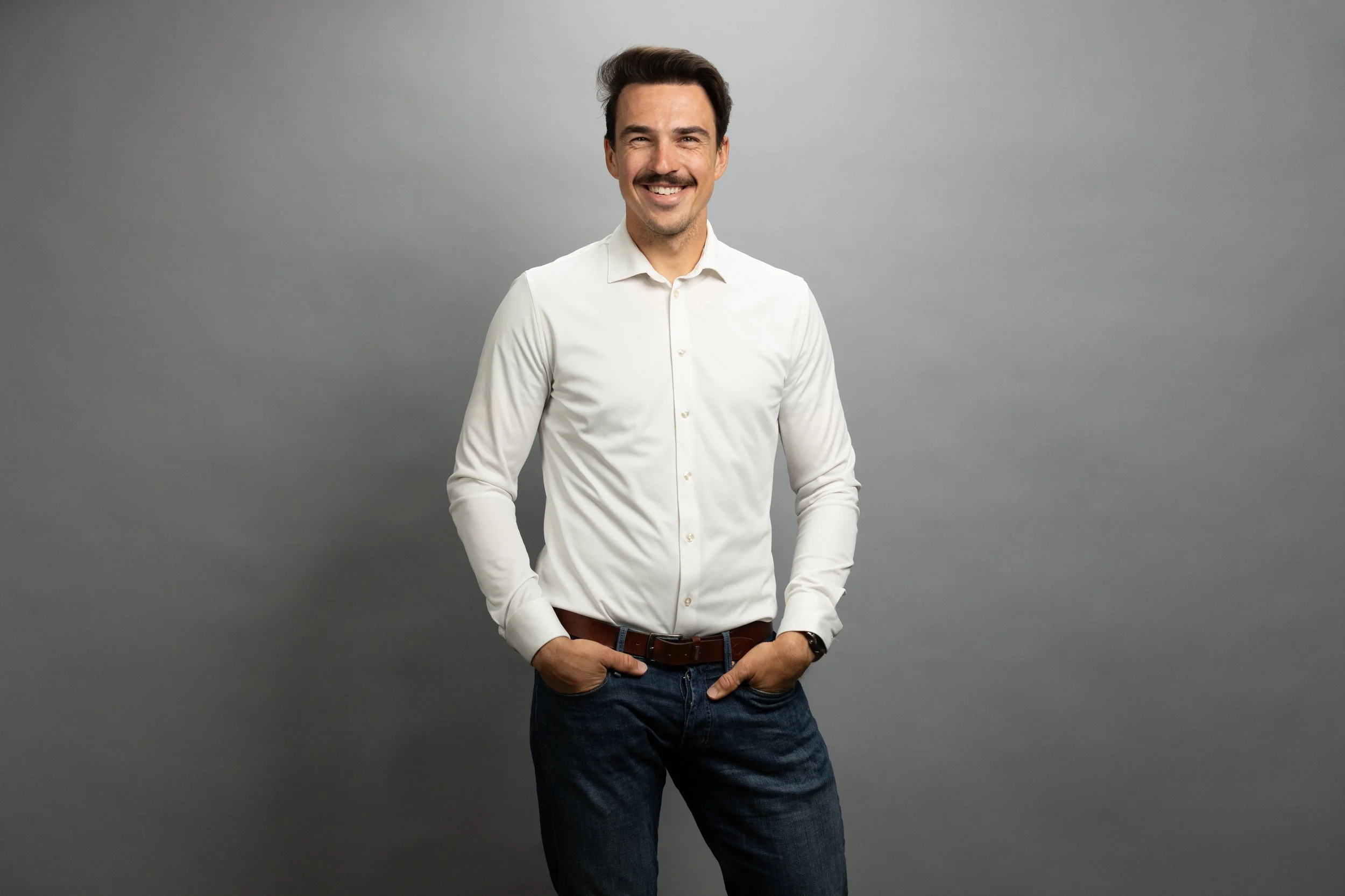 A man with dark hair and a mustache, wearing a white dress shirt and dark jeans, smiling and standing against a gray background.