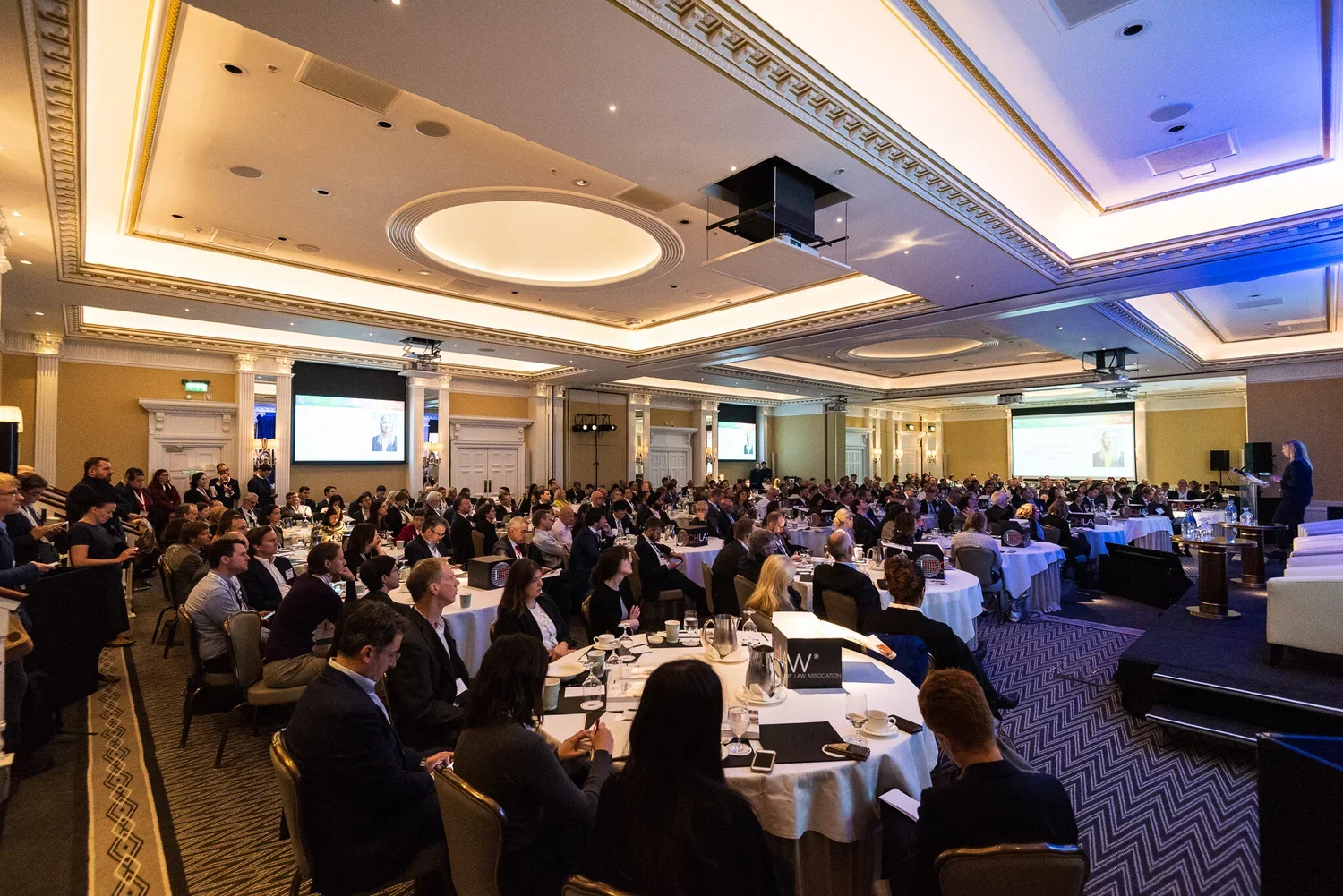 Large conference room filled with many attendees sitting at round tables, facing a speaker on stage. Two large screens display a presentation. The room has elegant lighting and decor.