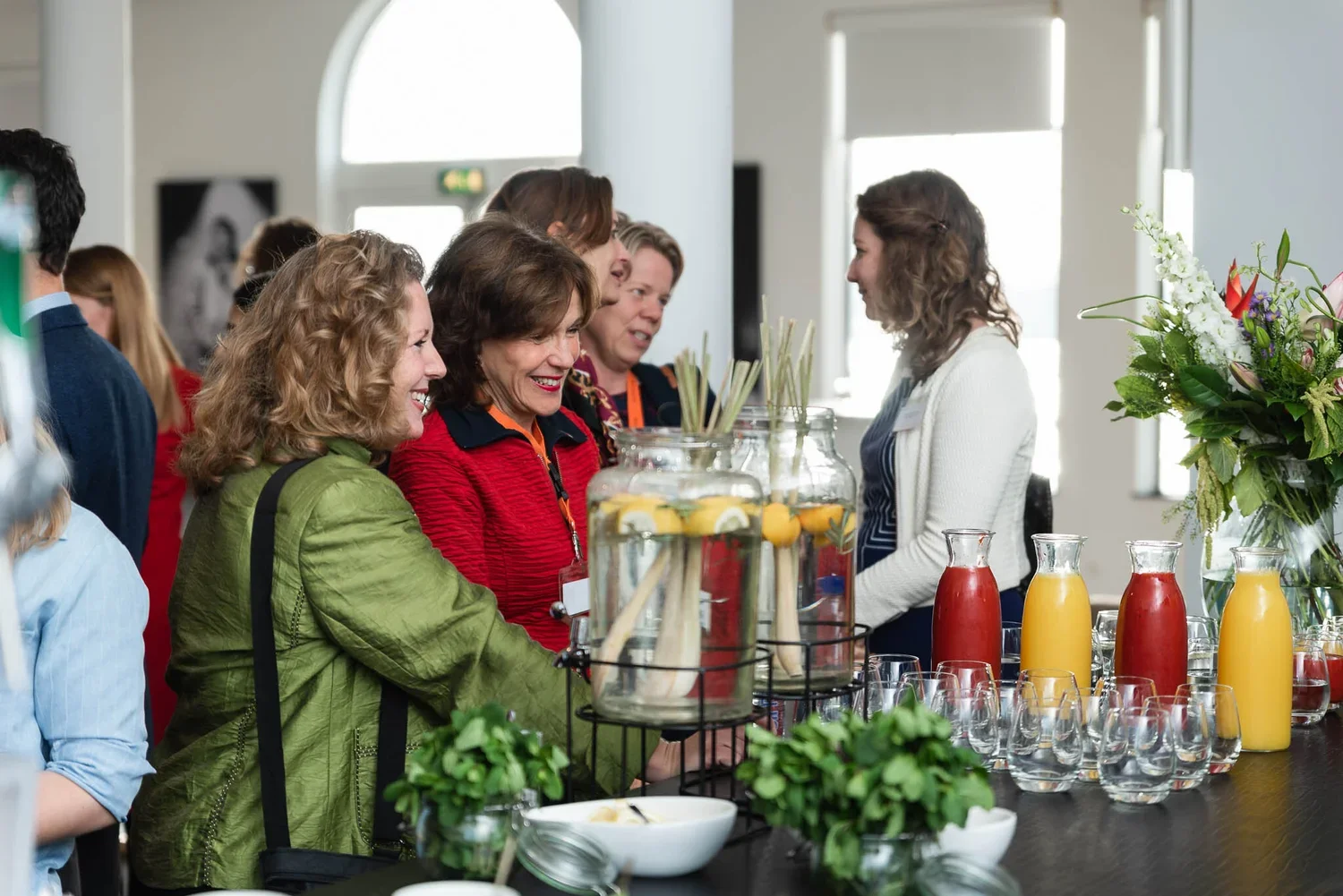 Women smiling and conversing at a food and drink station with pitchers of lemon water and juices, glasses, and a large flower arrangement in the background.