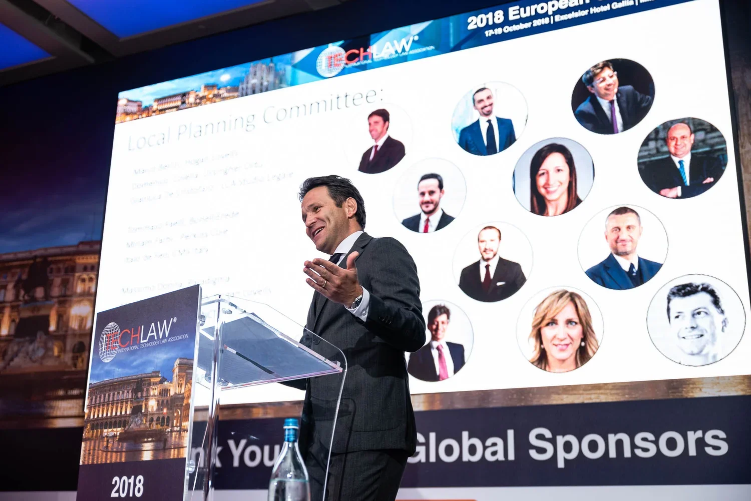 A man in a suit is speaking at a podium during a conference, with a large screen behind him displaying various headshots of people and the text '2018 European' and 'Local Planning Committee'. A water bottle is on the podium, and the background shows 