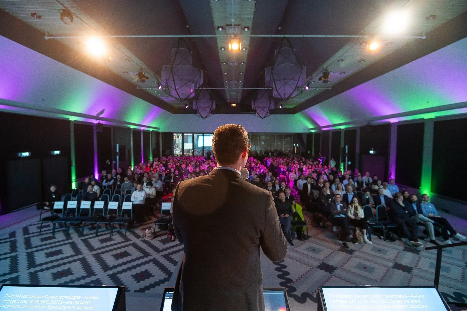 A man in a suit giving a presentation at a conference or seminar, facing a large audience seated in a hall with colorful lighting.
