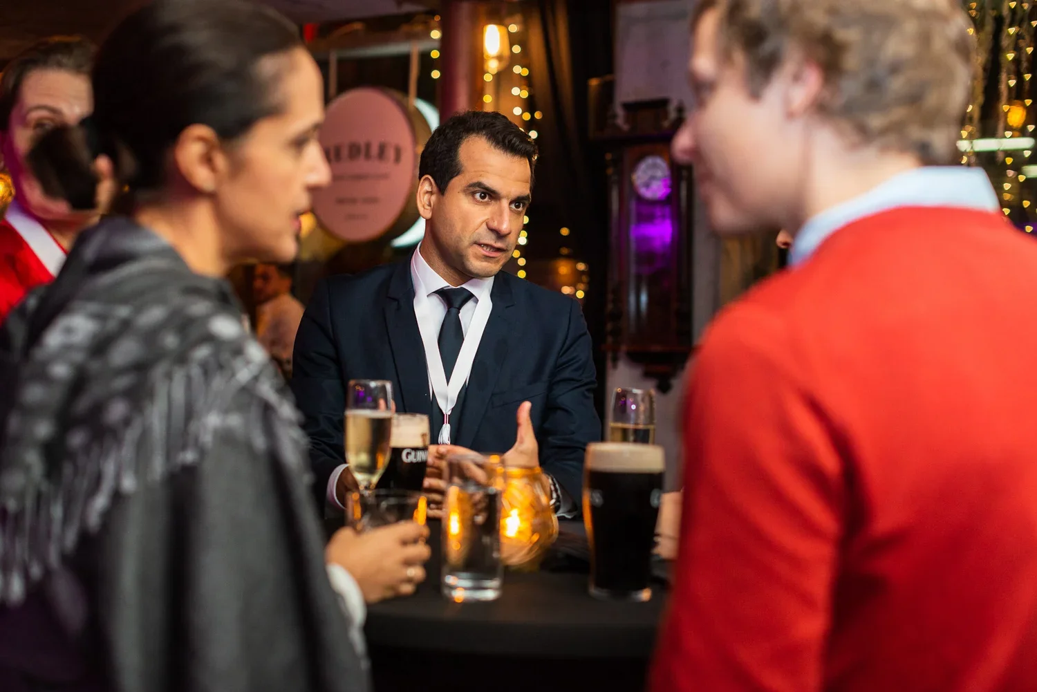 A man in a dark suit and tie is sitting at a bar with three other people, holding a glass of beer, engaging in conversation in a lively, decorated pub or bar setting.