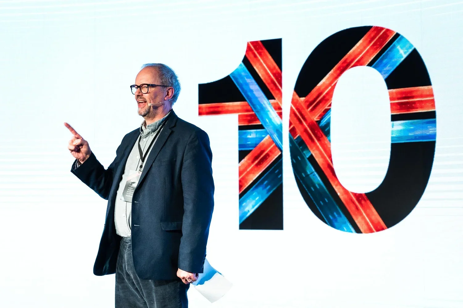 A man in glasses and a suit speaking on stage with a large '10' and colorful abstract design in the background.