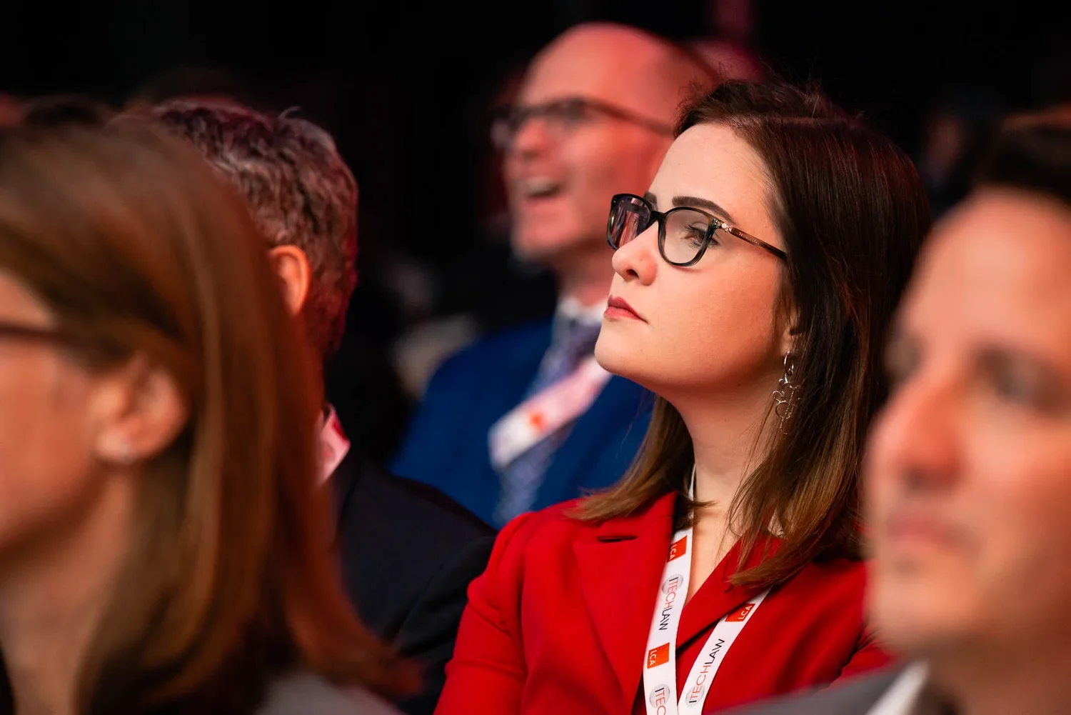 A woman wearing glasses and a red blazer, attending a conference, with other blurred attendees around her.