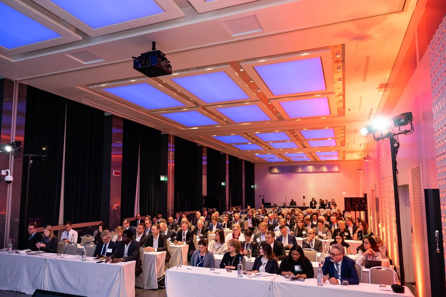 Conference room filled with attendees seated at long tables, with some people taking notes. The ceiling has large illuminated panels with blue and pink lighting, and there are stage lights on stands.