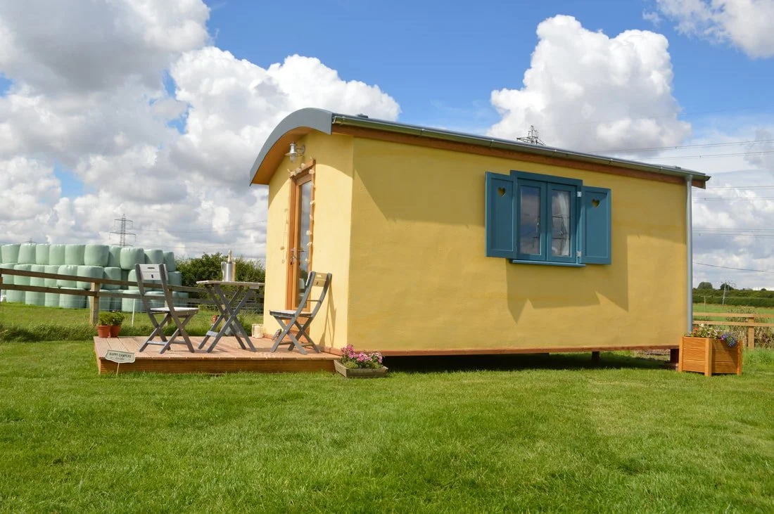 Yellow tiny house with blue window shutters on a grassy field, porch with table and chairs, cloudy sky.
