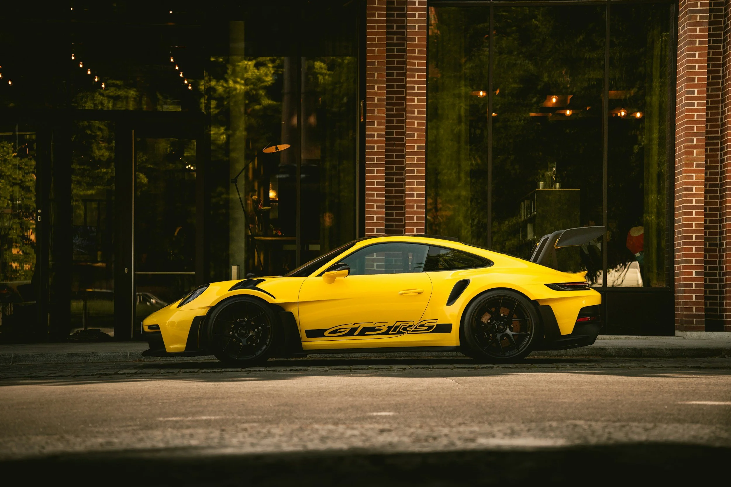 A yellow Porsche GT3 RS sports car parked on the street in front of a glass-fronted building with brick accents at dusk.