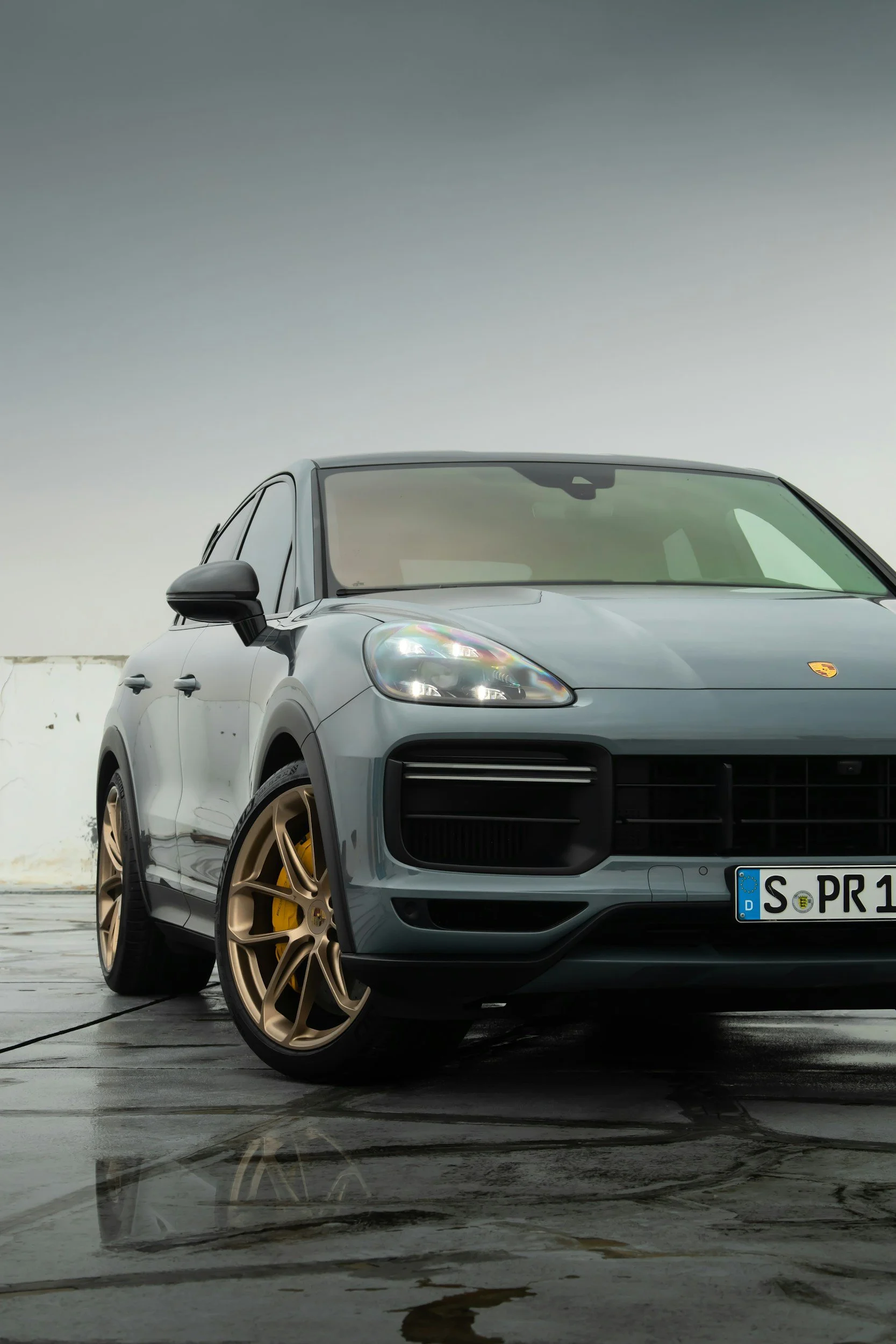 Gray Porsche SUV with gold wheels parked on wet surface against a cloudy sky.