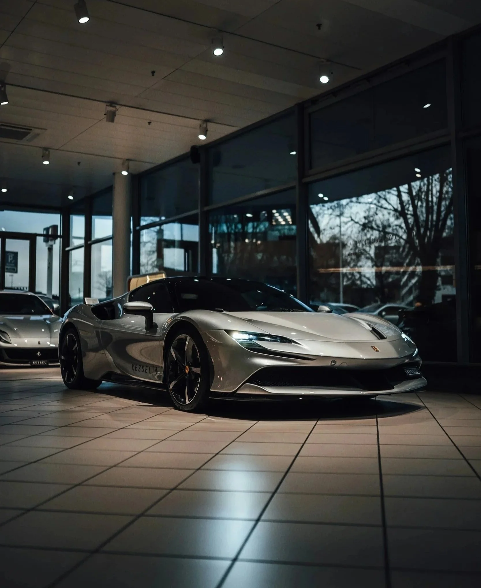 A silver sports car displayed inside a showroom with large glass windows showing trees outside, with tiled flooring and ceiling lights.