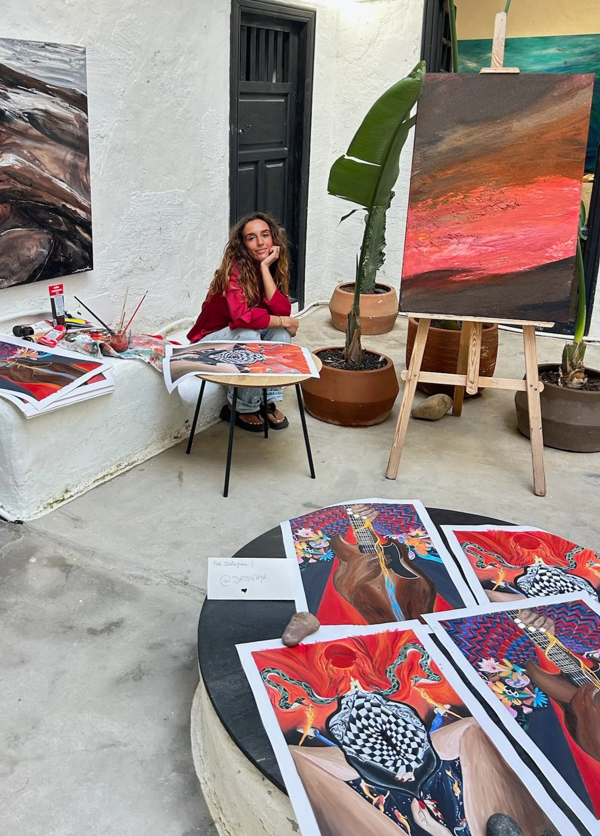 Une femme assise à côté de ses œuvres d'art peintes à la main, exposées sur une table, un chevalet, et autour d'elle plusieurs tableaux colorés en style abstrait. L'environnement semble être une galerie ou un espace d'exposition en extérieur.