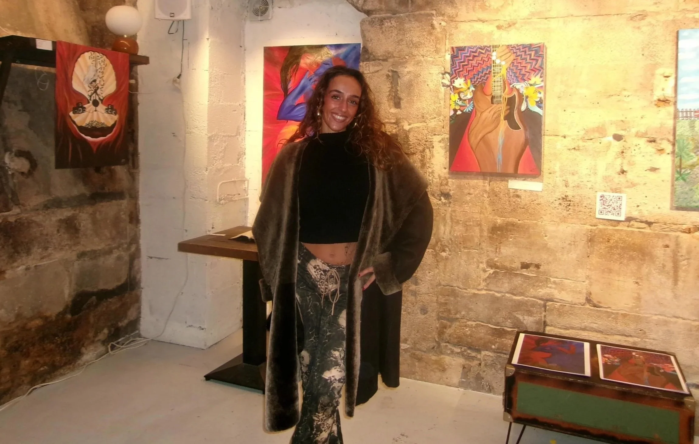 Une femme souriante se tient dans une galerie d'art avec des œuvres colorées en arrière-plan, y compris des peintures abstraites représentant des figures humaines et un guitariste. Le mur en pierre et l'ambiance chaleureuse complètent la scène.