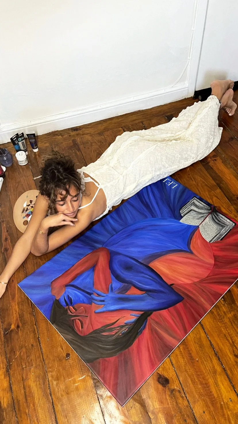 Femme peignant un tableau de style abstrait avec des couleurs vives comme le rouge, le bleu et le noir, allongée sur un sol en bois, portant une robe blanche, avec une palette de peinture à côté.