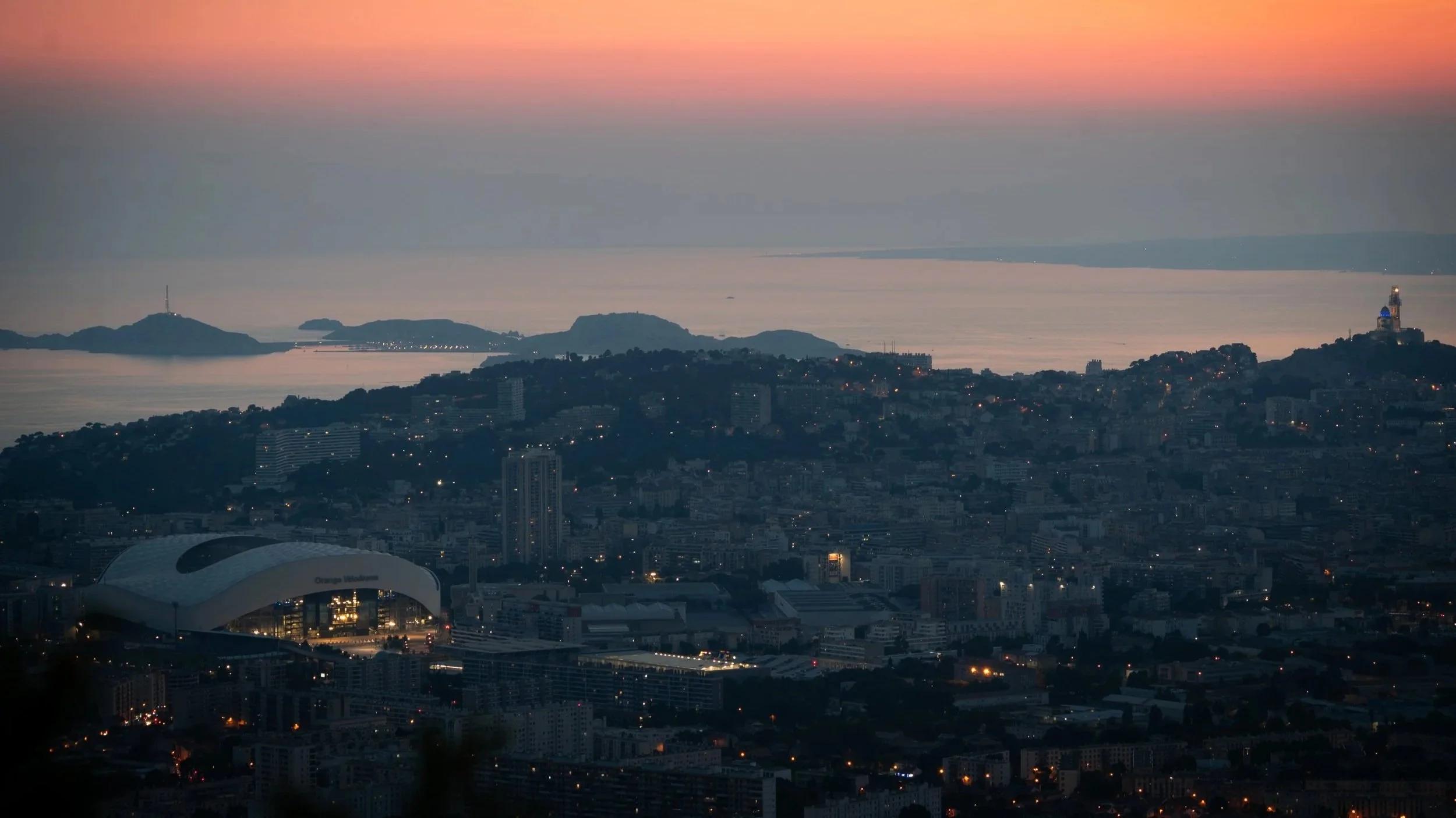 Vue panoramique de marseille au coucher du soleil avec le stade vélodrome en premier plan. Le ciel présente des couleurs chaudes allant du rose à l'orange.