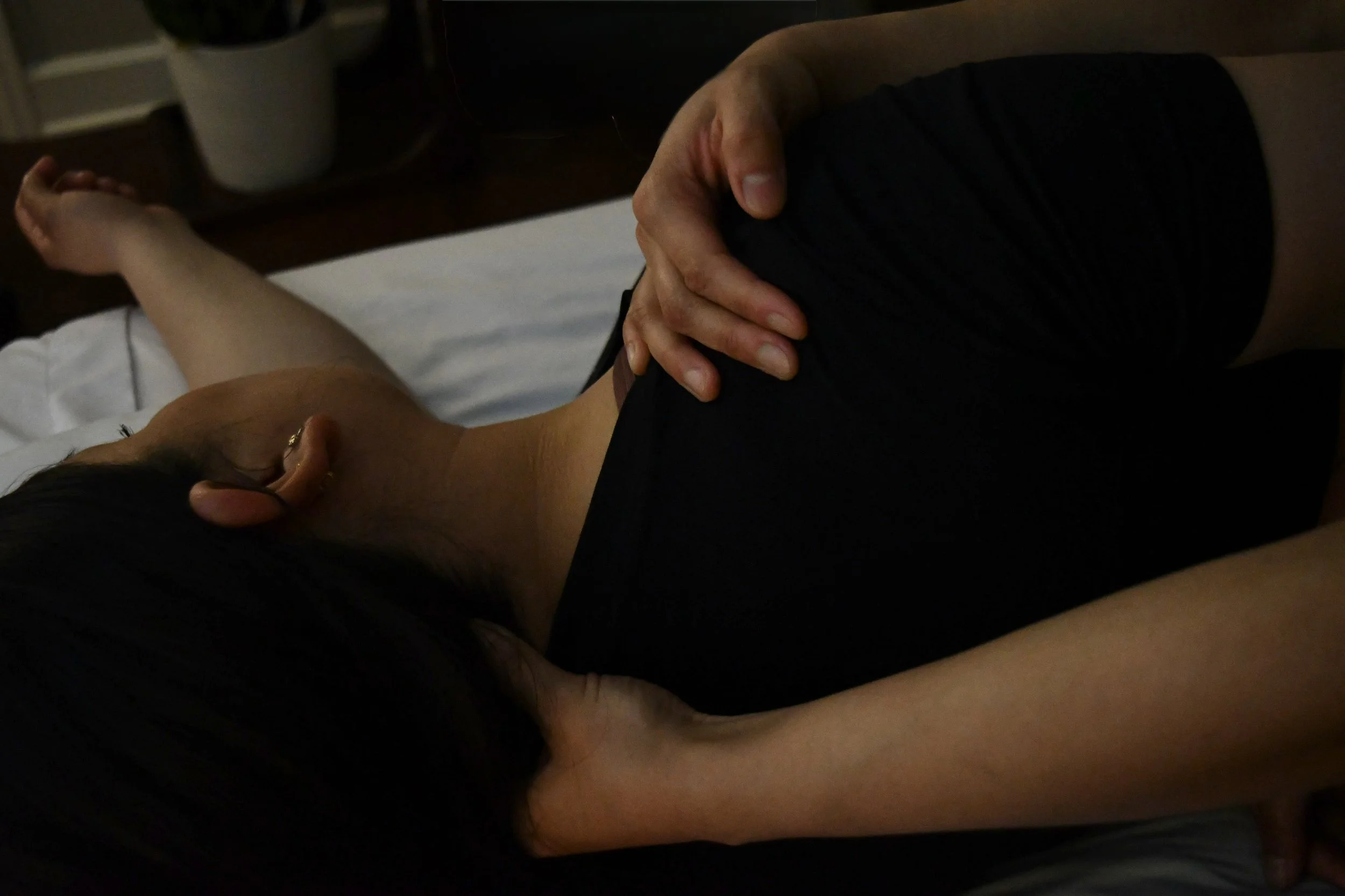 A person lying on their side on a bed, with one hand on their stomach. They are dressed in black clothing.