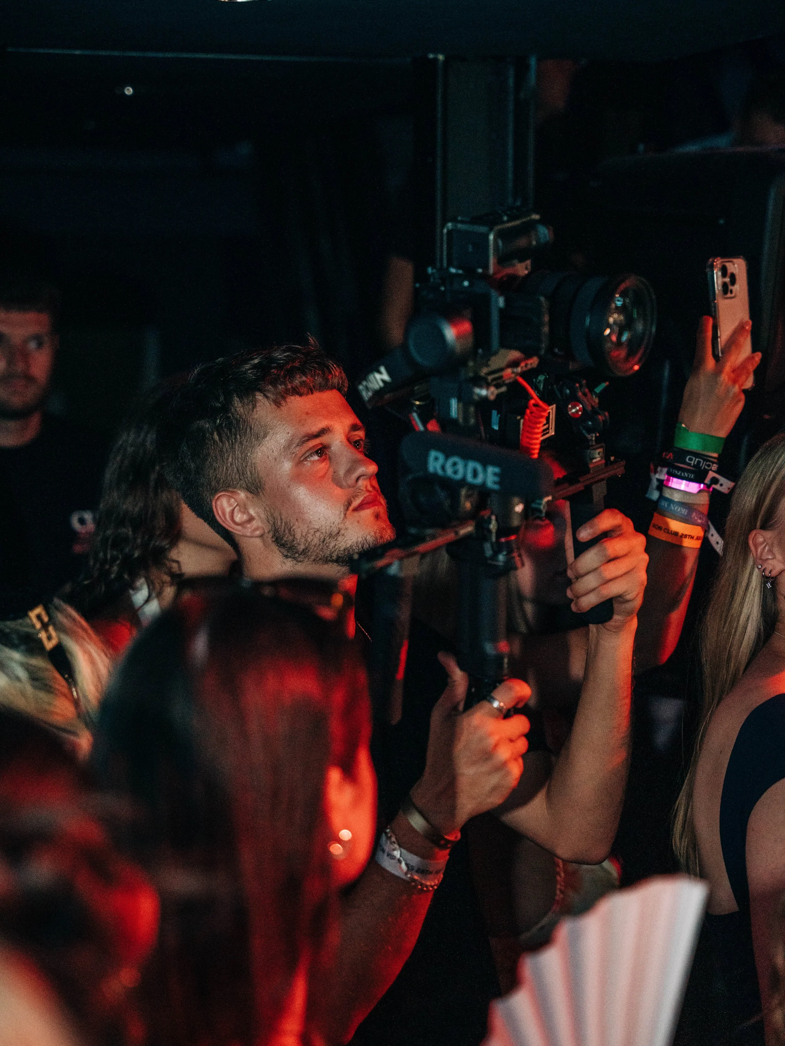 A man holding a video camera in a crowded, dark event with colorful lighting.