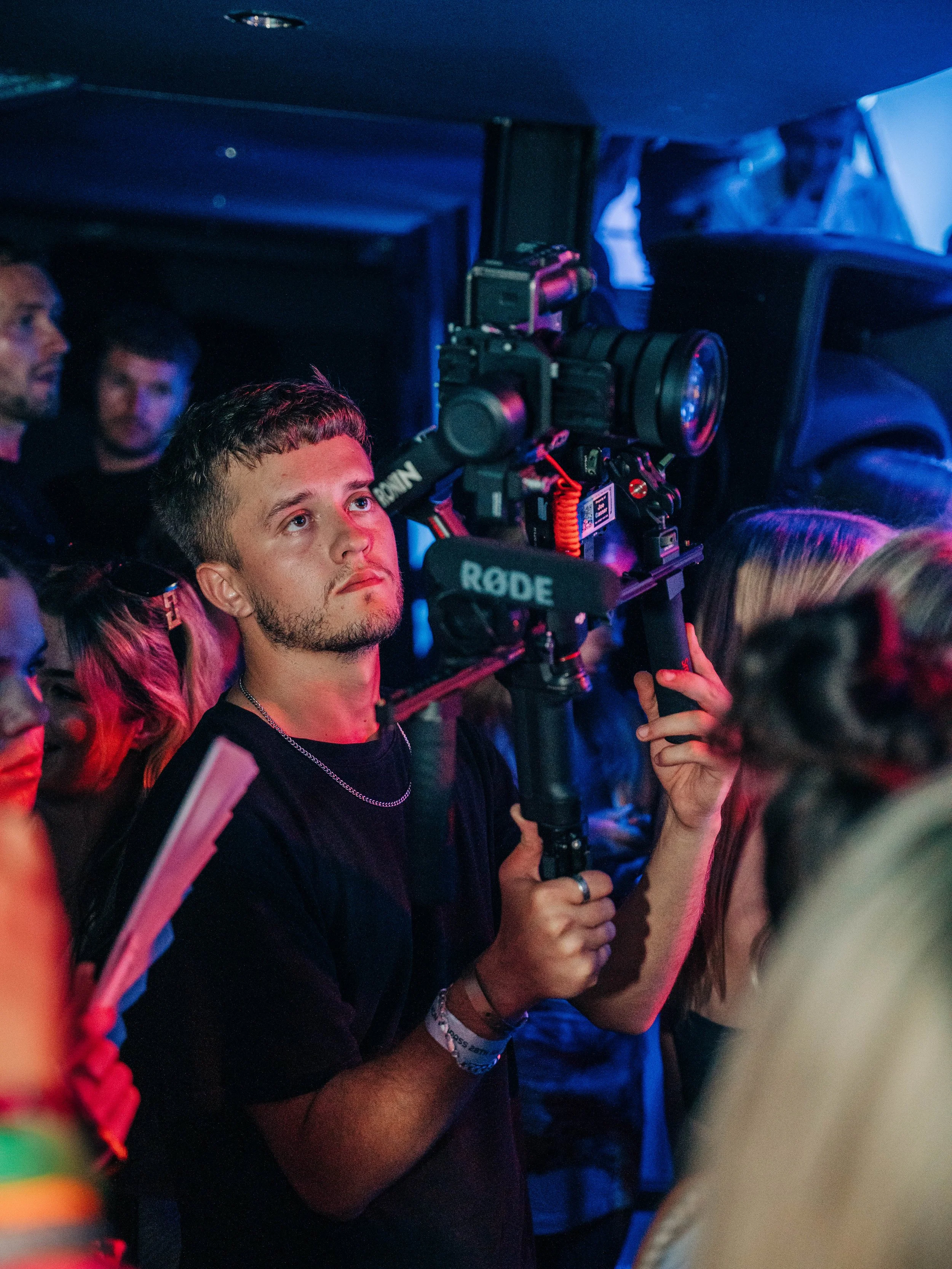 A young man operating a professional video camera at an event, surrounded by other attendees under colorful lighting.