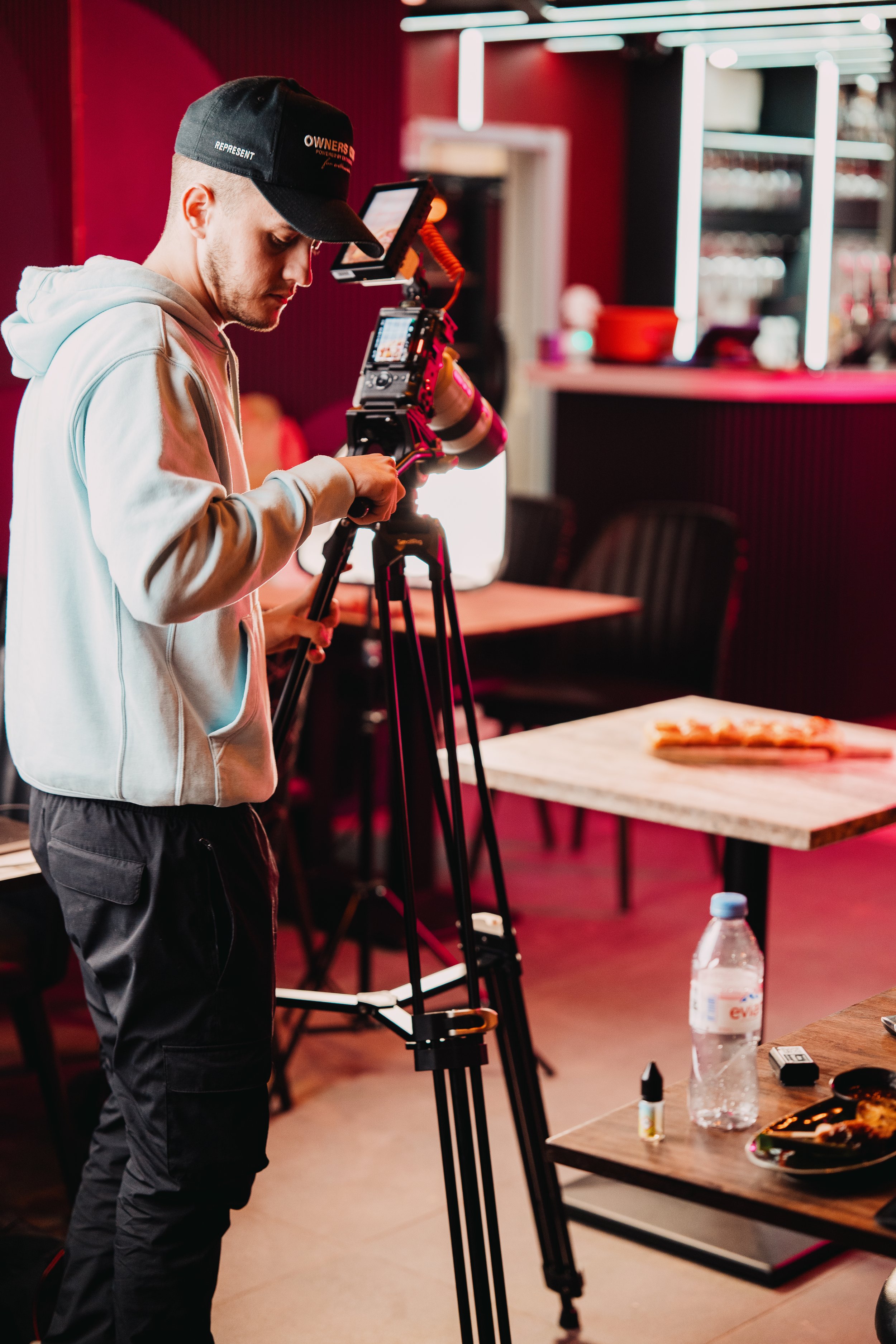 A videographer operating a professional camera on a tripod inside a restaurant or cafe with dark red walls, black chairs, and tables. There is a water bottle, a plate, and small items on a table in the foreground.