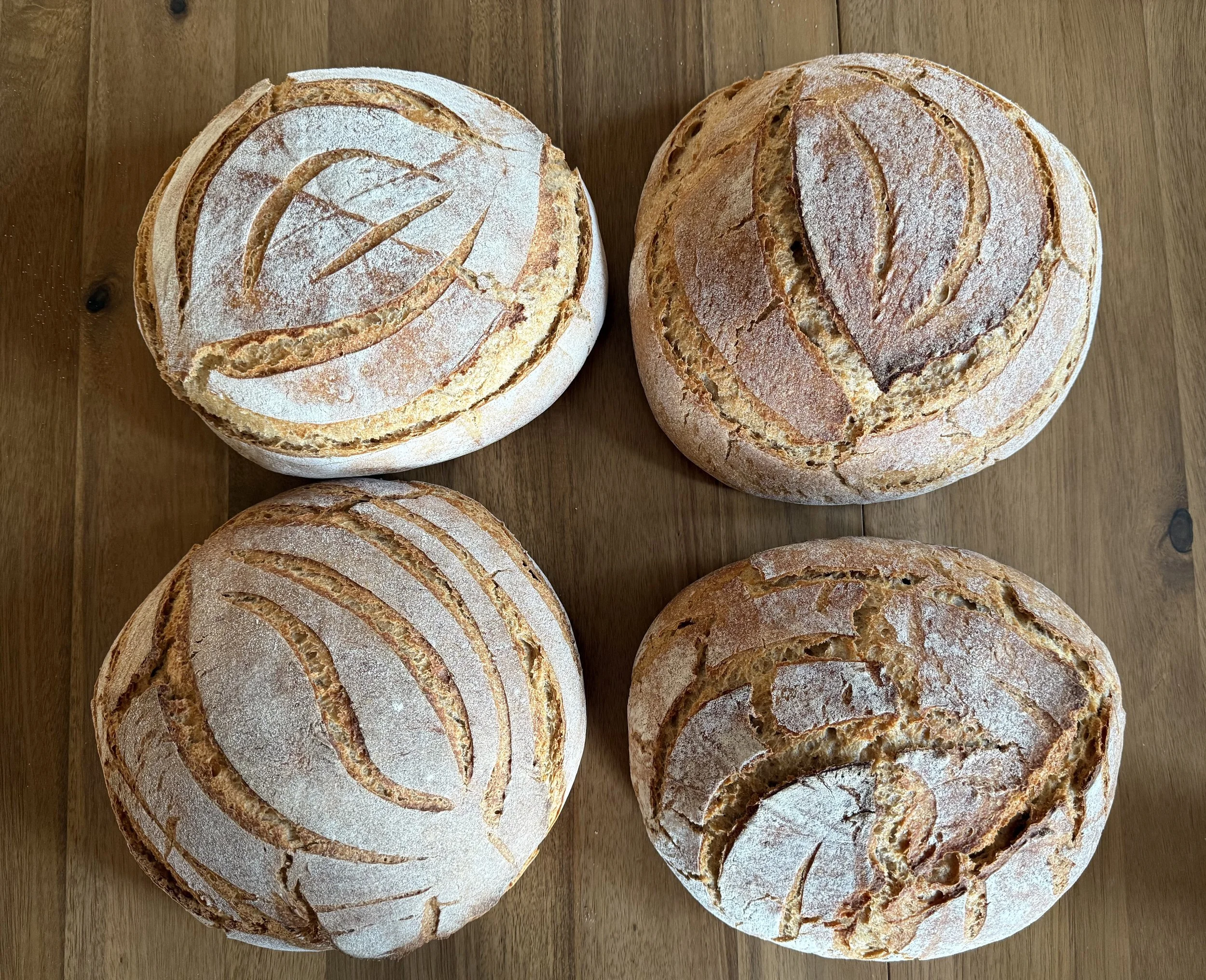 Four rounds of freshly baked artisanal bread with patterns scored into the crust, placed on a wooden surface.