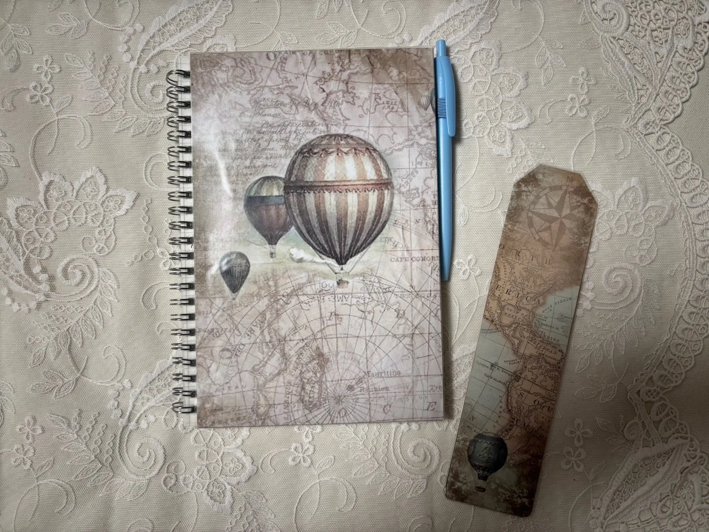 Notizbuch mit Ballonmotiven, blauer Kugelschreiber und eine Lesezeichen mit Weltkarten-Motiv auf einem Tischtuch mit Blumenmuster.