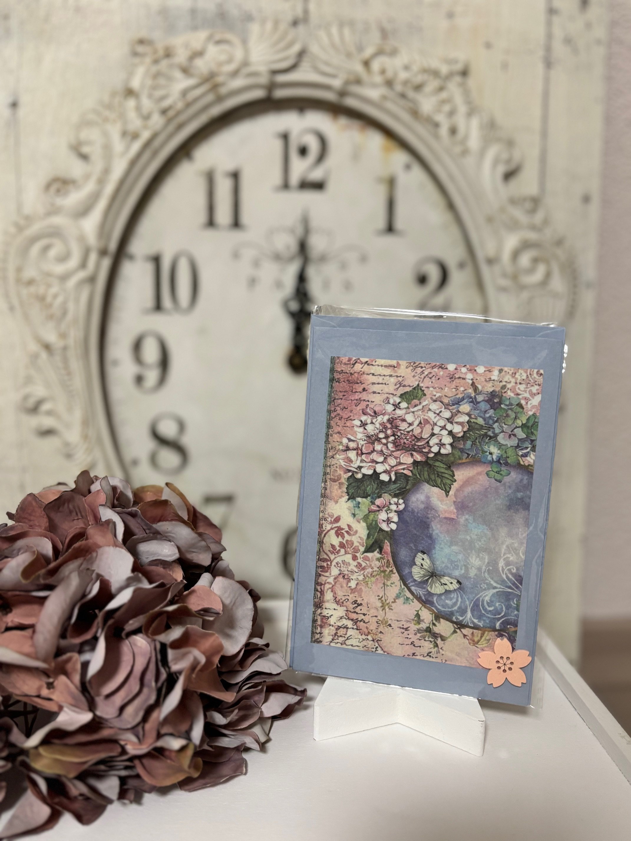 Eine goldene Wanduhr zeigt 12:00 Uhr. Vor der Uhr stehen beige und pinkfarbene Papierblumen und eine Grußkarte mit einer Blumenvase, Schmetterling und Blumenmuster.