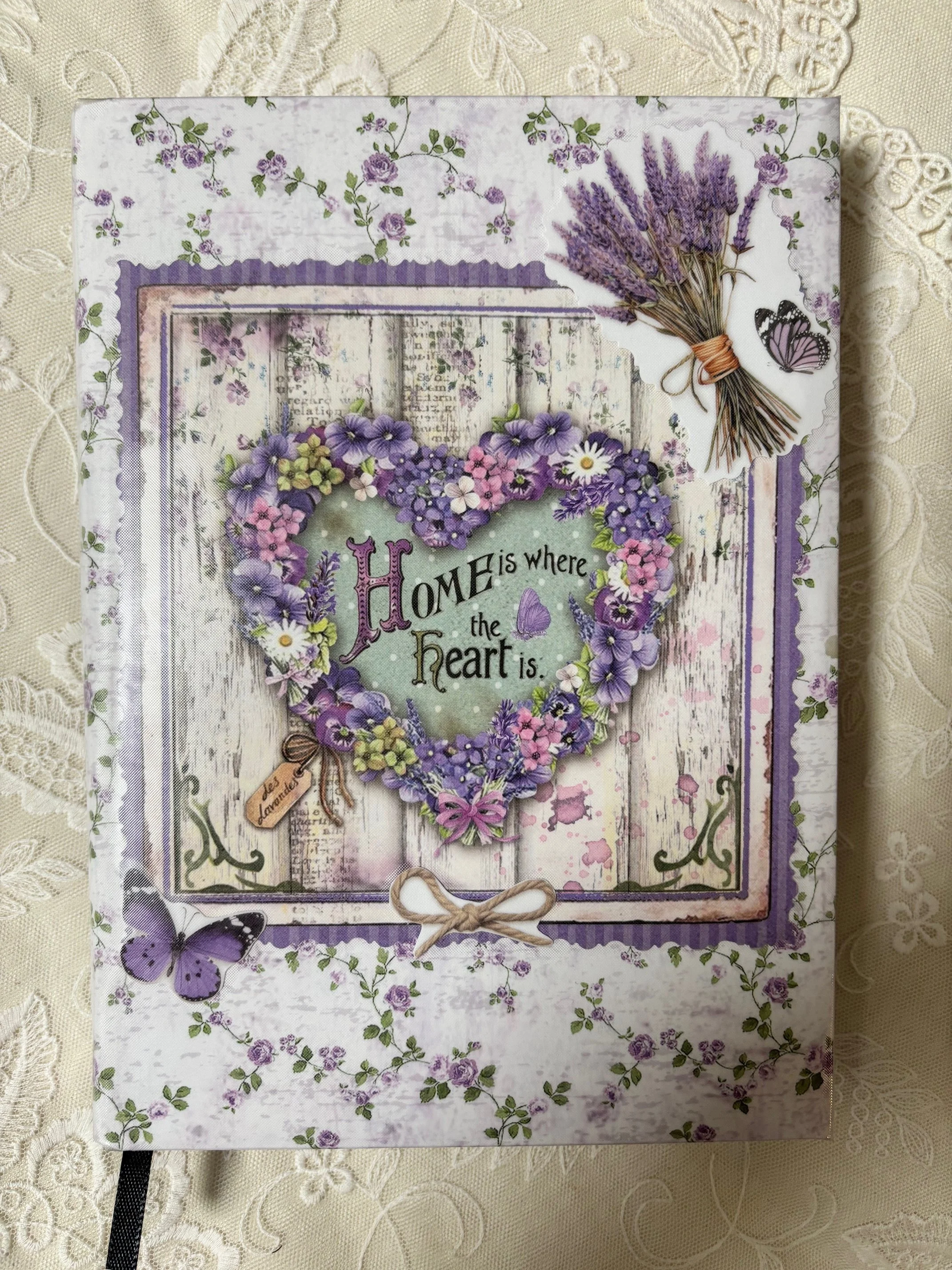 Ein dekoratives Buchcover mit einem Herz aus Lavendel und Blumen. Der Text lautet "Home is where the heart is". Es gibt auch Butterfly- und Lavendel-Accessoires auf dem Cover.