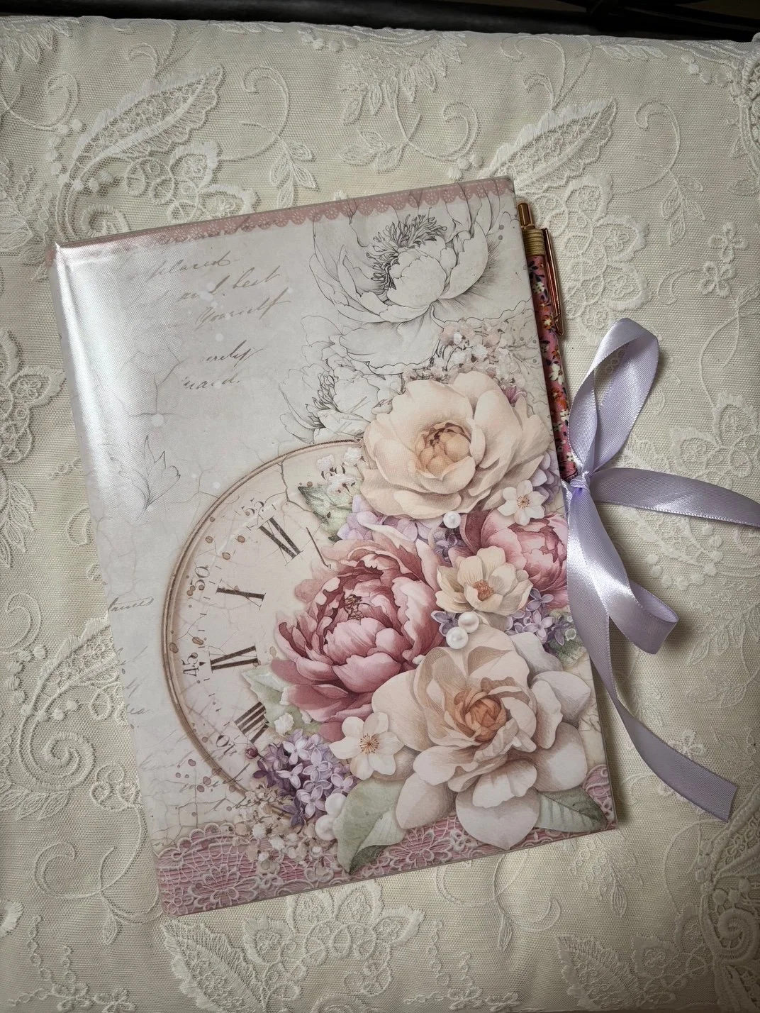 Ein wunderschön gestaltetes Notizbuch mit Blumenmuster, einer Uhr und einem Satinband auf einem Stickerei-Tuch.