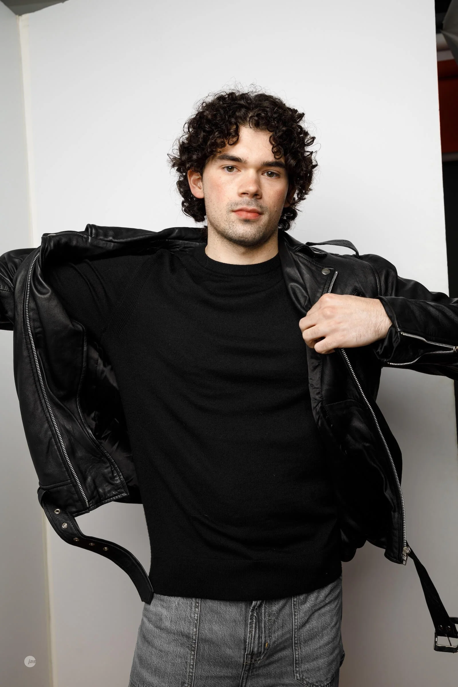A young man with curly dark hair, wearing a black leather jacket over a black shirt, and gray pants, standing against a plain white background.