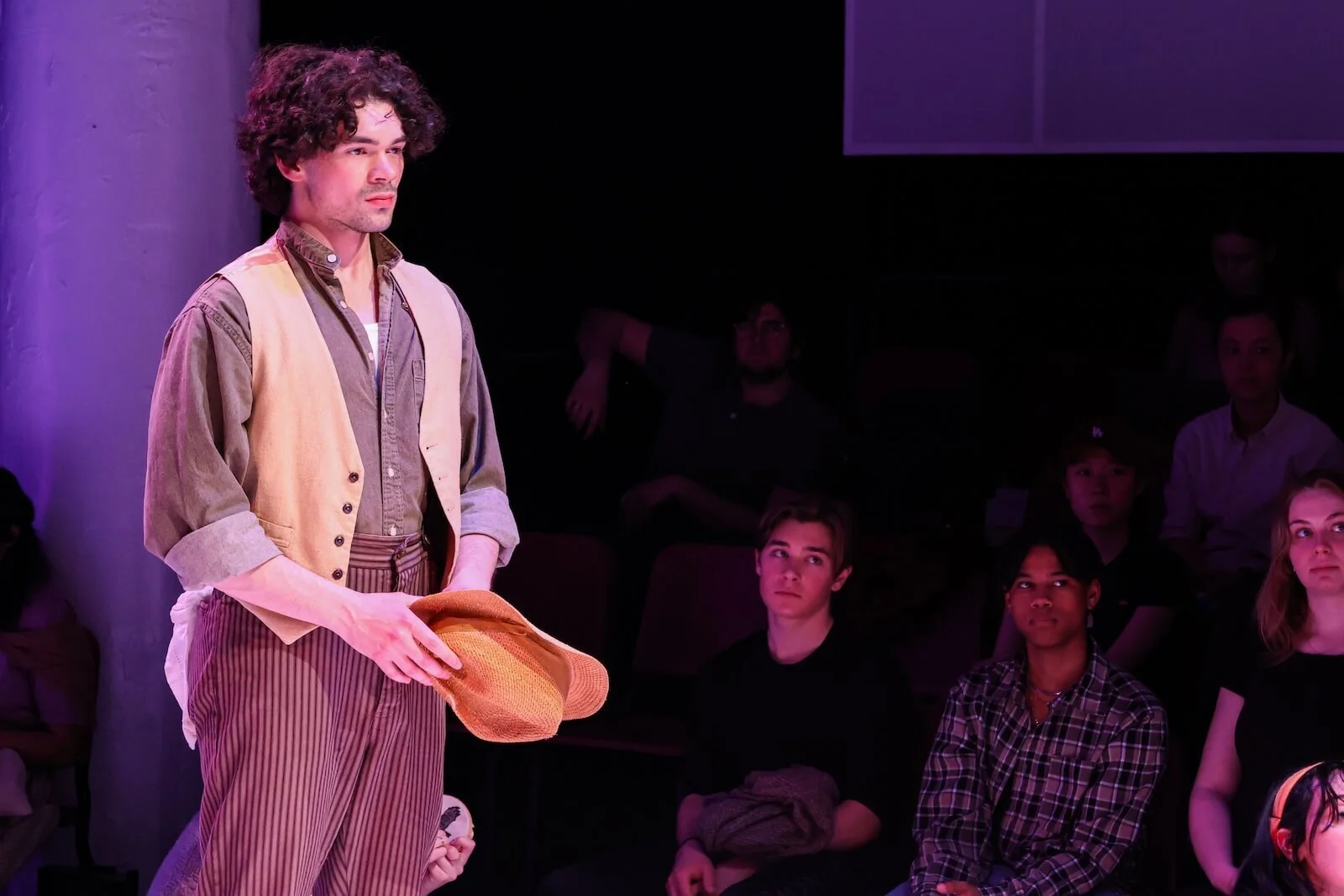 A young man with curly hair in vintage clothing standing on stage holding a straw hat, with an audience watching him in the background.